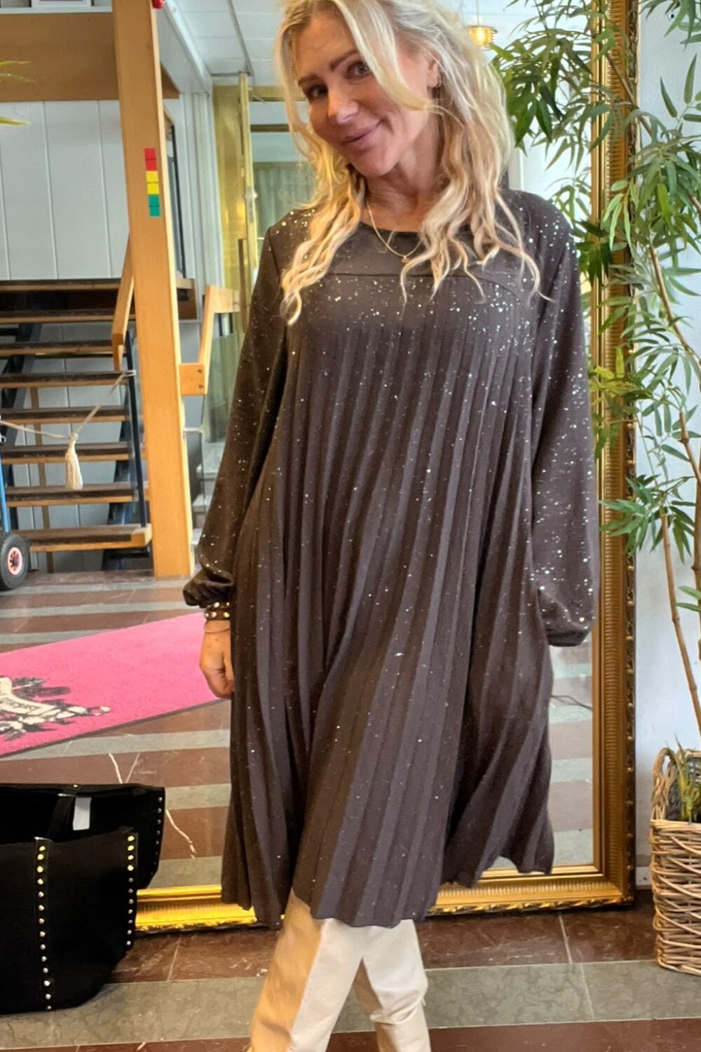 Leende blond kvinna poserar framför en stor guldramsspegel inomhus, med huvudet lätt på sned. Hon bär en mörk glittrig, plisserad klänning och vita stövlar. I bakgrunden syns trappa, växter och inramade foton.