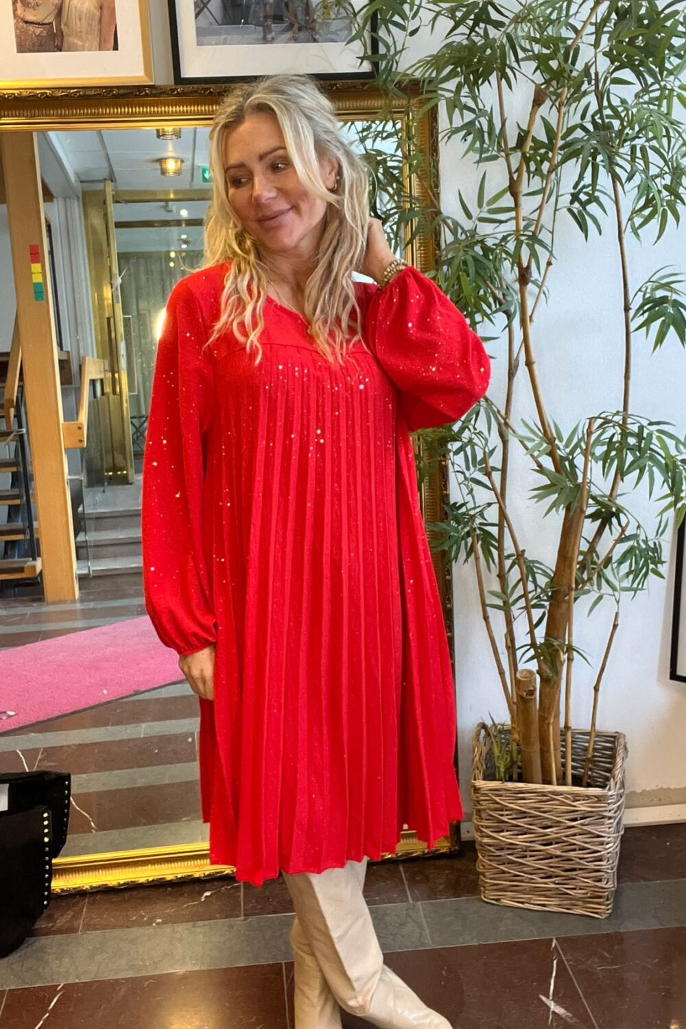 Blond kvinna poserar med handen i håret och korsade ben, iklädd röd glittrig, veckad klänning och vita höga stövlar. Hon står inomhus vid en stor guldramsspegel, växt i korg och inramade foton på väggen.
