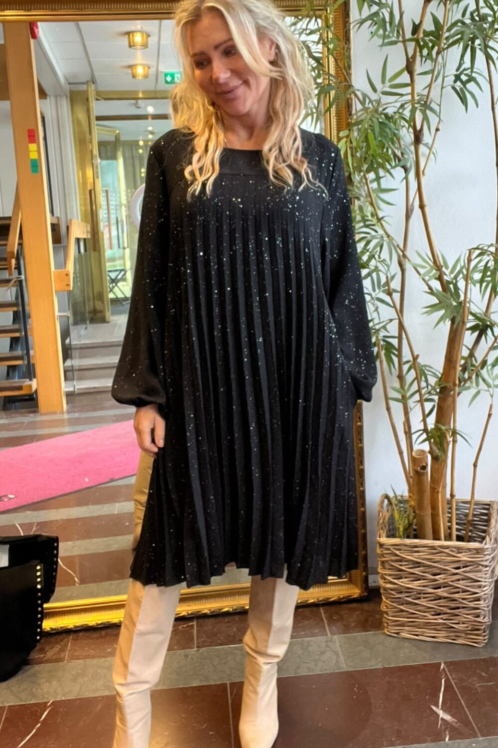Blond kvinna står och poserar framför en stor guldramsspegel i en inomhusmiljö, klädd i svart glittrig plisserad klänning och beige stövlar. Hon ler lätt; bredvid syns en hög krukväxt och en korridor.