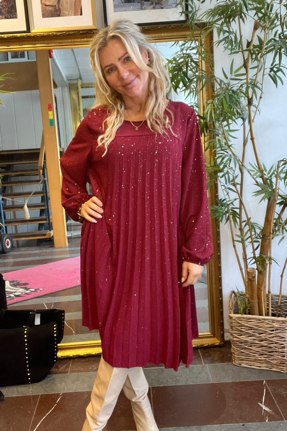 Leende kvinna poserar med handen på höften i en vinröd, glittrig långärmad klänning och ljusa boots. Hon står inomhus framför en stor guldramsspegel, med en hög bambuväxt och inramade foton på väggen.