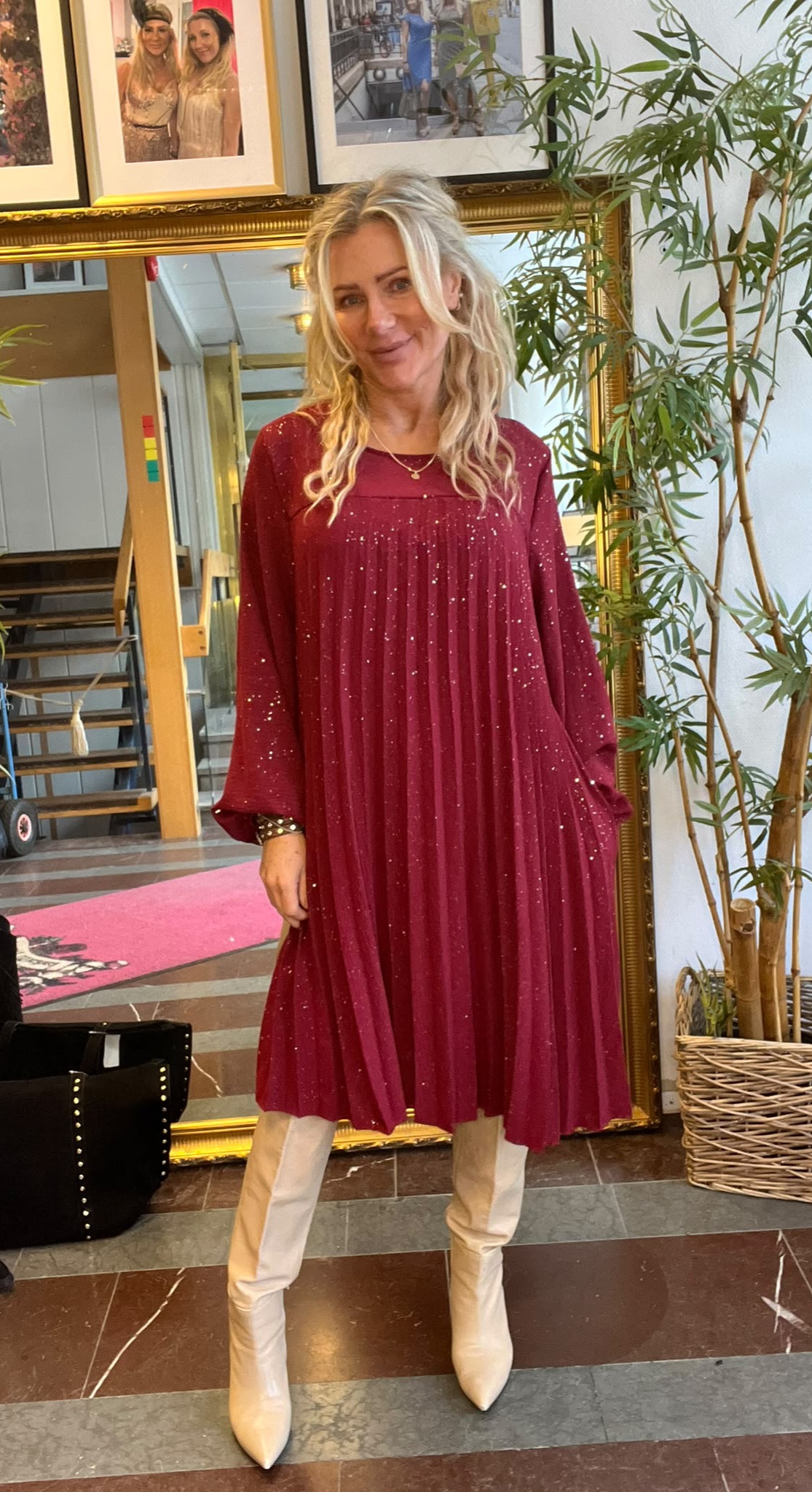 Blond kvinna står och ler framför en stor guldramsspegel, klädd i vinröd glittrig klänning med vida ärmar och vita knähöga stövlar. Inomhusmiljö med trappa, bambuliknande växt och inramade foton ovanför spegeln.