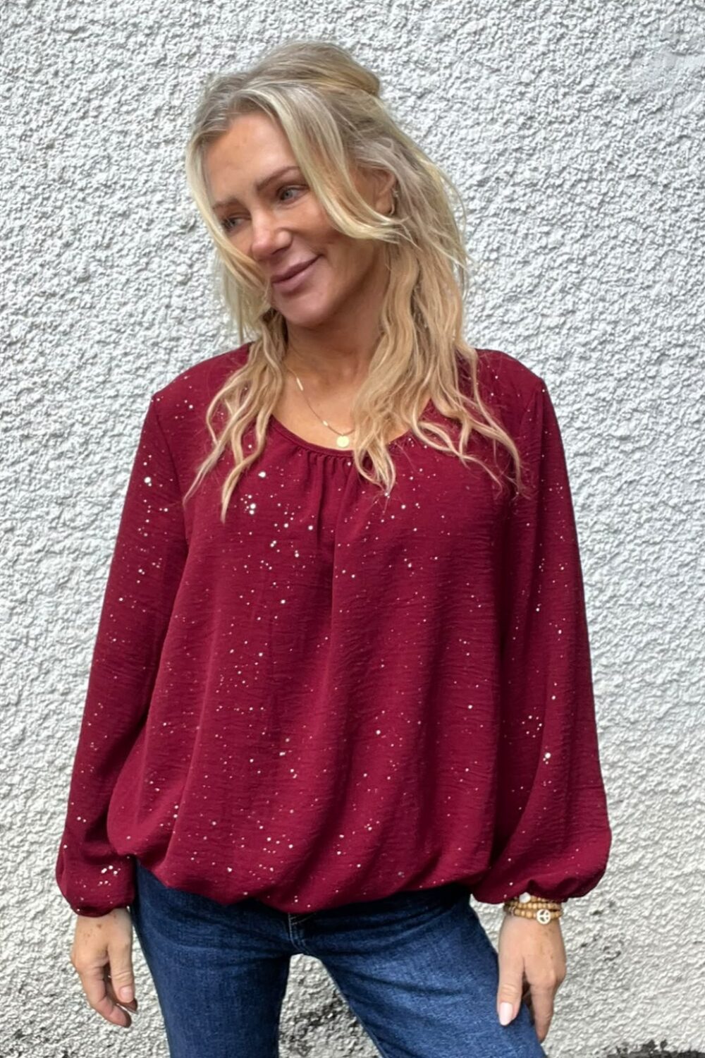 Blond kvinna står avslappnat mot en ljus, grovputsad vägg och tittar åt sidan med ett svagt leende. Hon bär vinröd blus med vita prickar, blå jeans och smycken på handled och hals.
