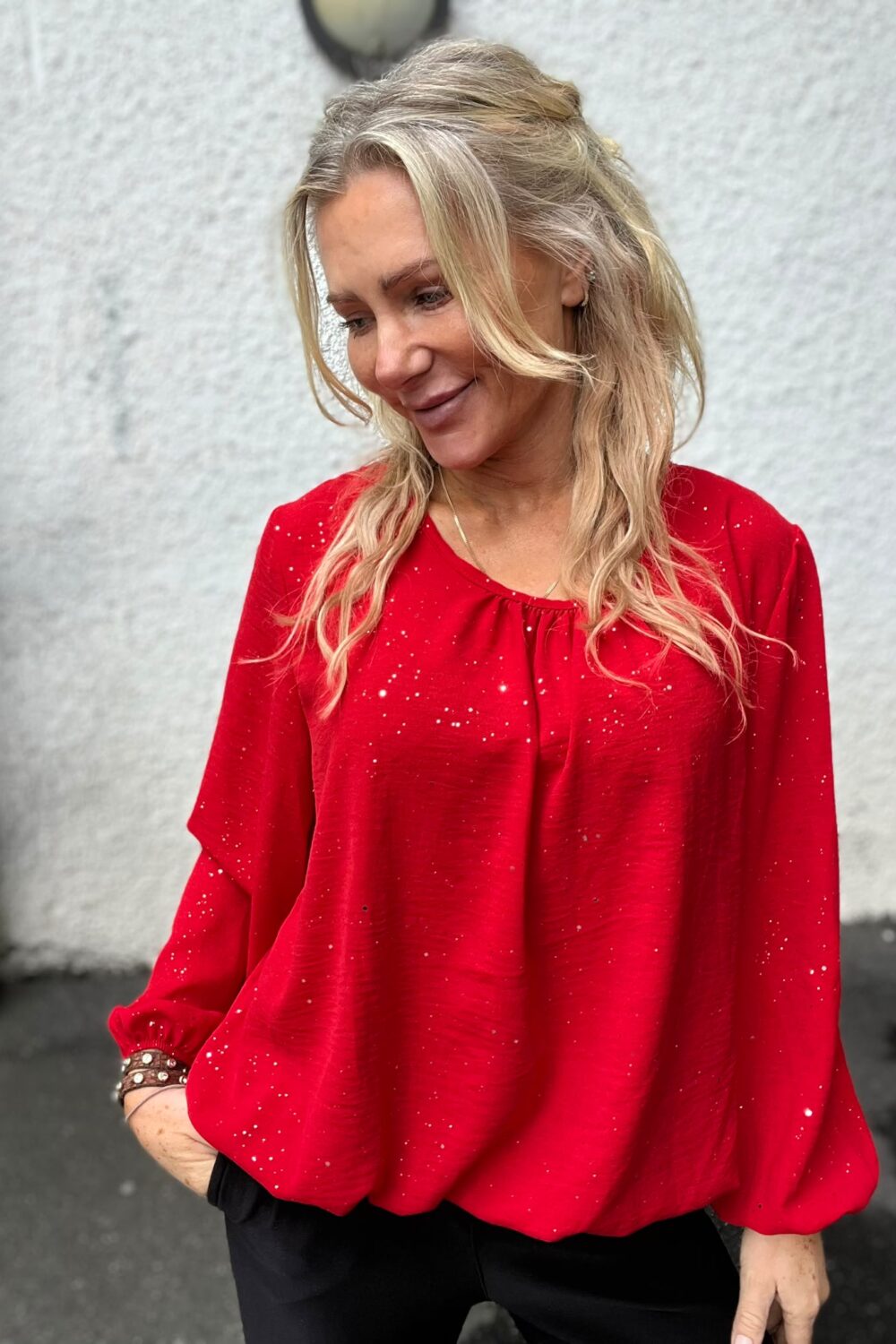 Blond kvinna i röd, glittrig blus står utomhus mot en ljus, strukturerad vägg och ler lätt medan hon tittar ned åt sidan. Hon bär armband och svarta byxor; bilden är ett halvkroppsporträtt.