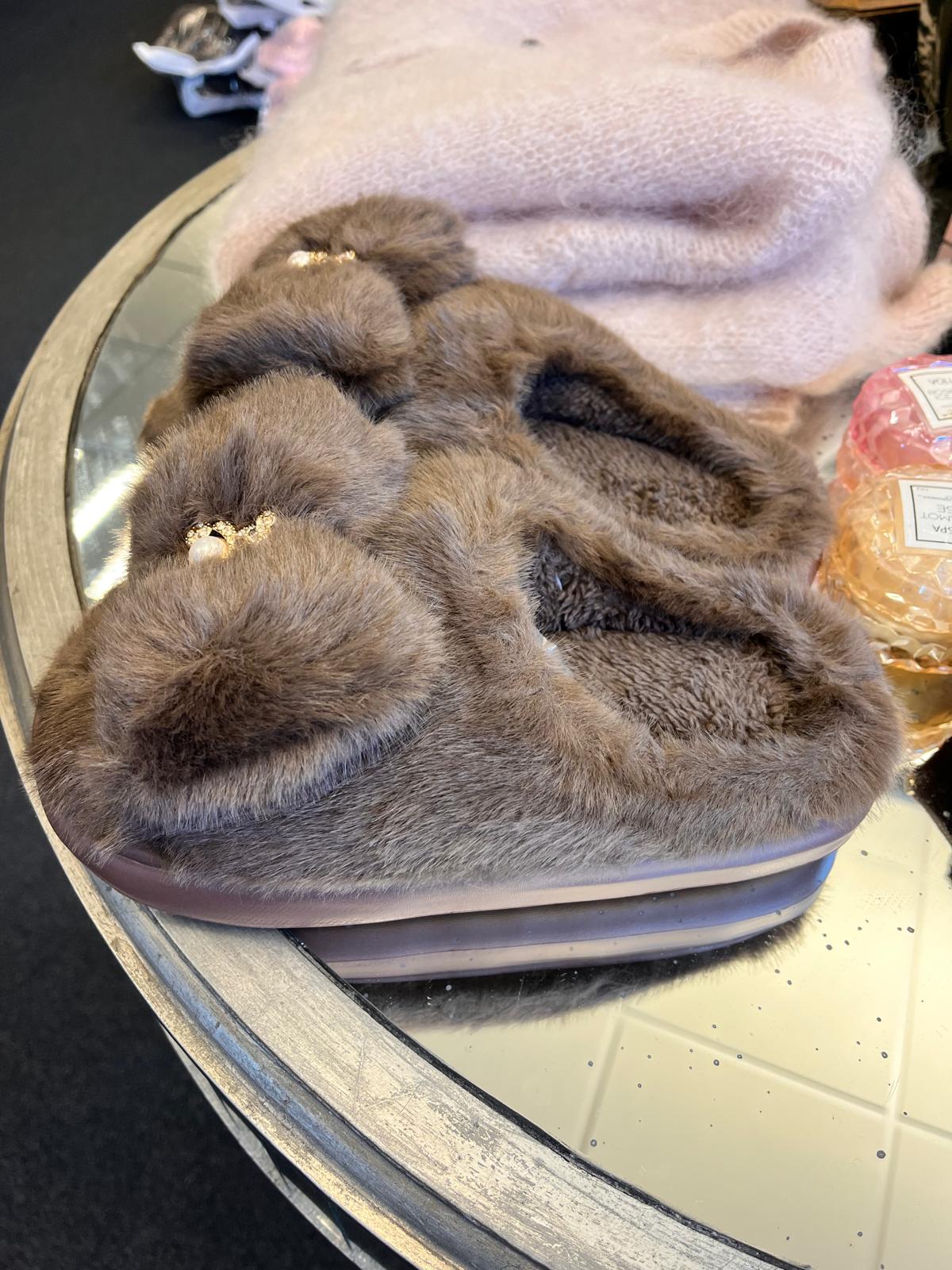 Grå, lurvig toffel med korsade band och ett litet pärl- och guldfärgat smycke ligger på ett runt spegelbord. I bakgrunden syns en ljusrosa stickad mössa och en rosa parfymflaska.