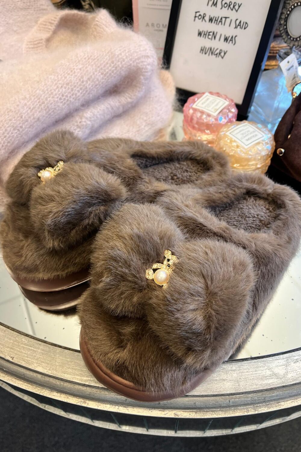 Bruna, fluffiga tofflor med pärl- och strassdetalj ligger på ett runt bord i en butiksmiljö, framför en ljusrosa mössa och två doftljus. I bakgrunden står en skylt: “I’M SORRY FOR WHAT I SAID WHEN I WAS HUNGRY”.