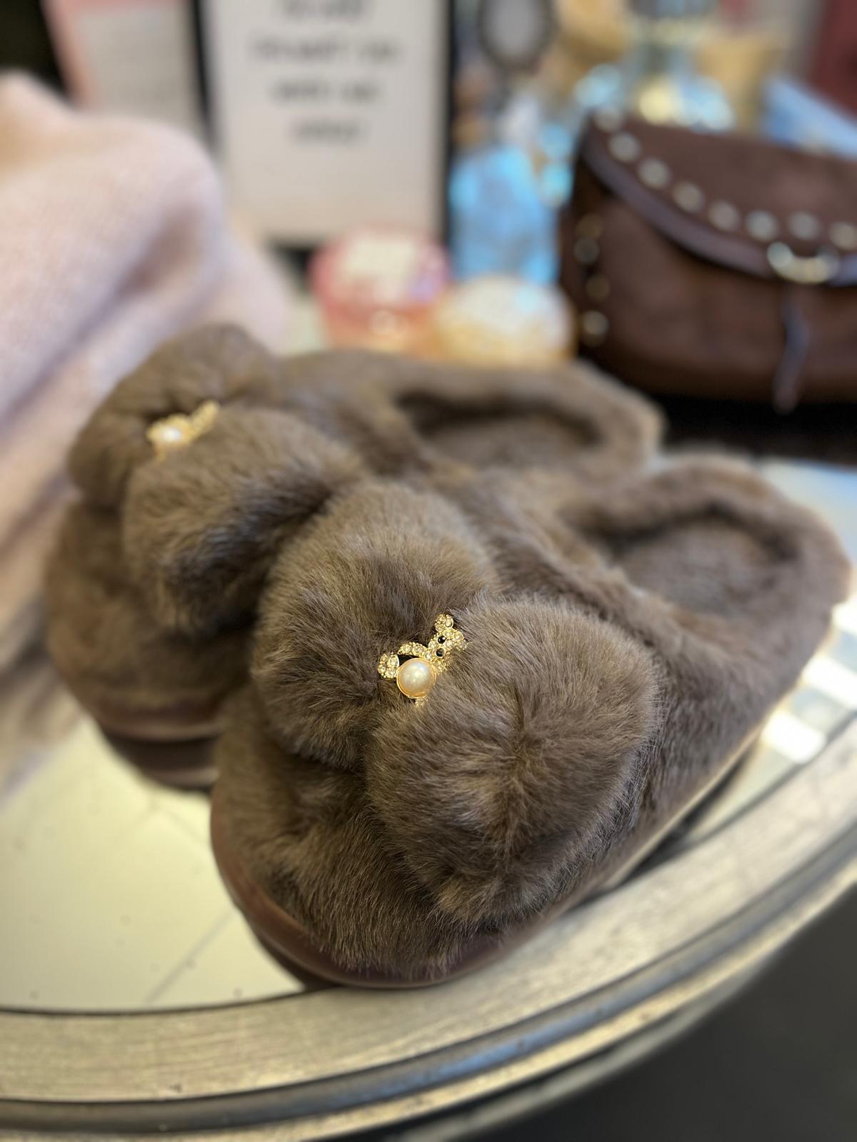 Ett par grå, fluffiga tofflor ligger på en rund spegelyta. Varje toffla har en liten guldfärgad brosch med pärla och strass framtill. I bakgrunden syns suddiga accessoarer, bland annat en brun väska.