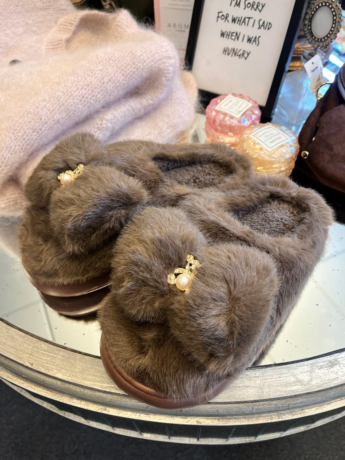 Bruna, fluffiga tofflor med pärl- och strassdetalj ligger på ett runt bord i en butiksmiljö, framför en ljusrosa mössa och två doftljus. I bakgrunden står en skylt: “I’M SORRY FOR WHAT I SAID WHEN I WAS HUNGRY”.