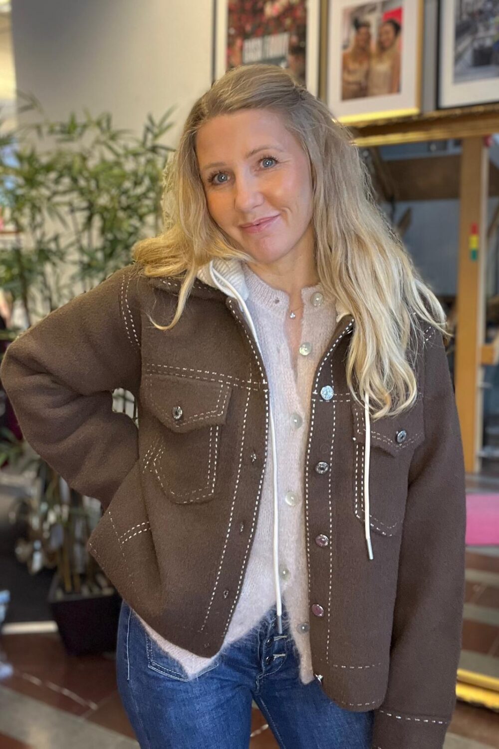 Blond kvinna står och poserar med handen på höften och ler mot kameran, klädd i brun jacka med vita sömmar över ljus kofta och jeans. I bakgrunden syns växter och inramade foton på väggen.