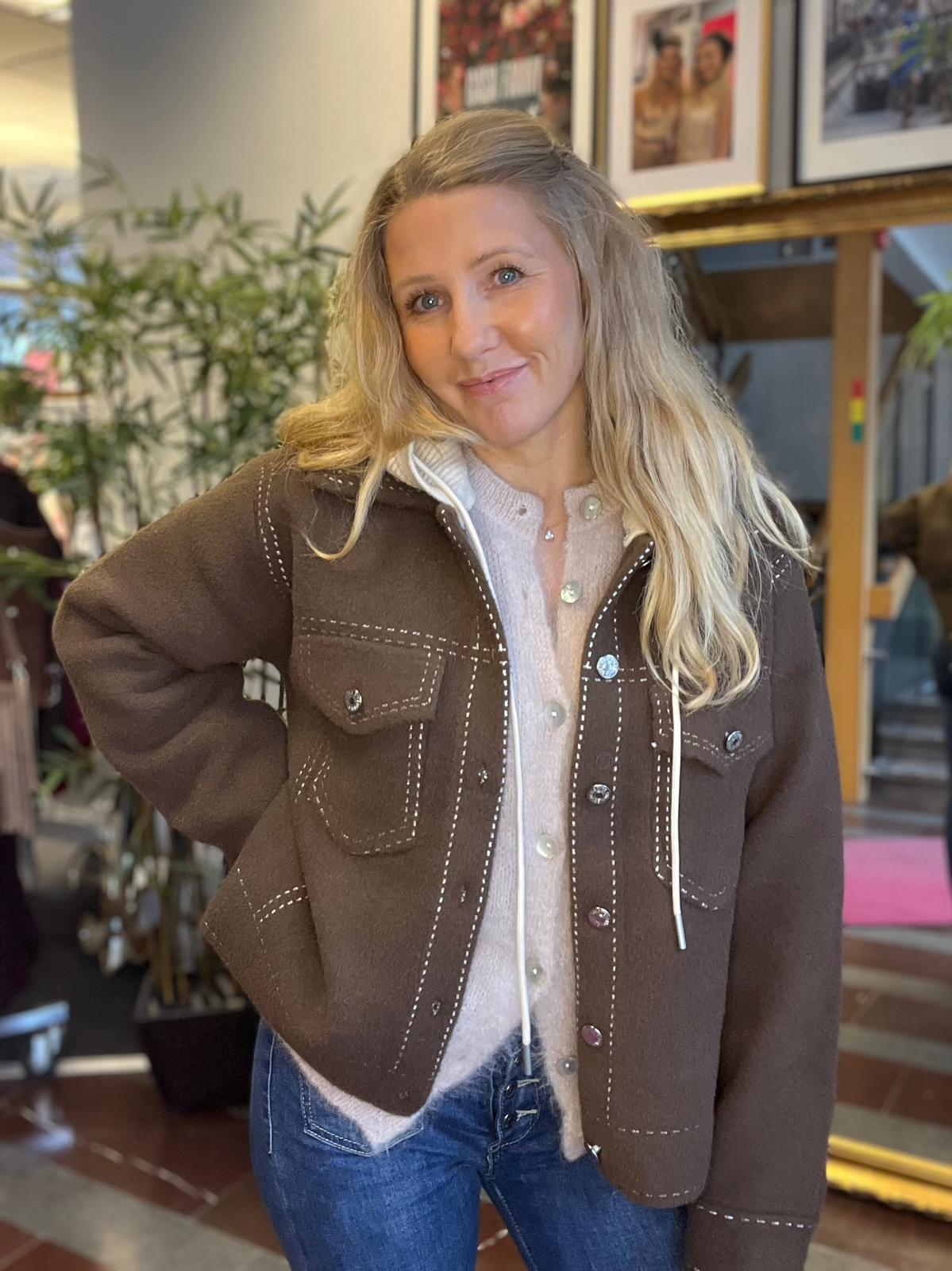 Blond kvinna står och poserar med handen på höften och ler mot kameran, klädd i brun jacka med vita sömmar över ljus kofta och jeans. I bakgrunden syns växter och inramade foton på väggen.