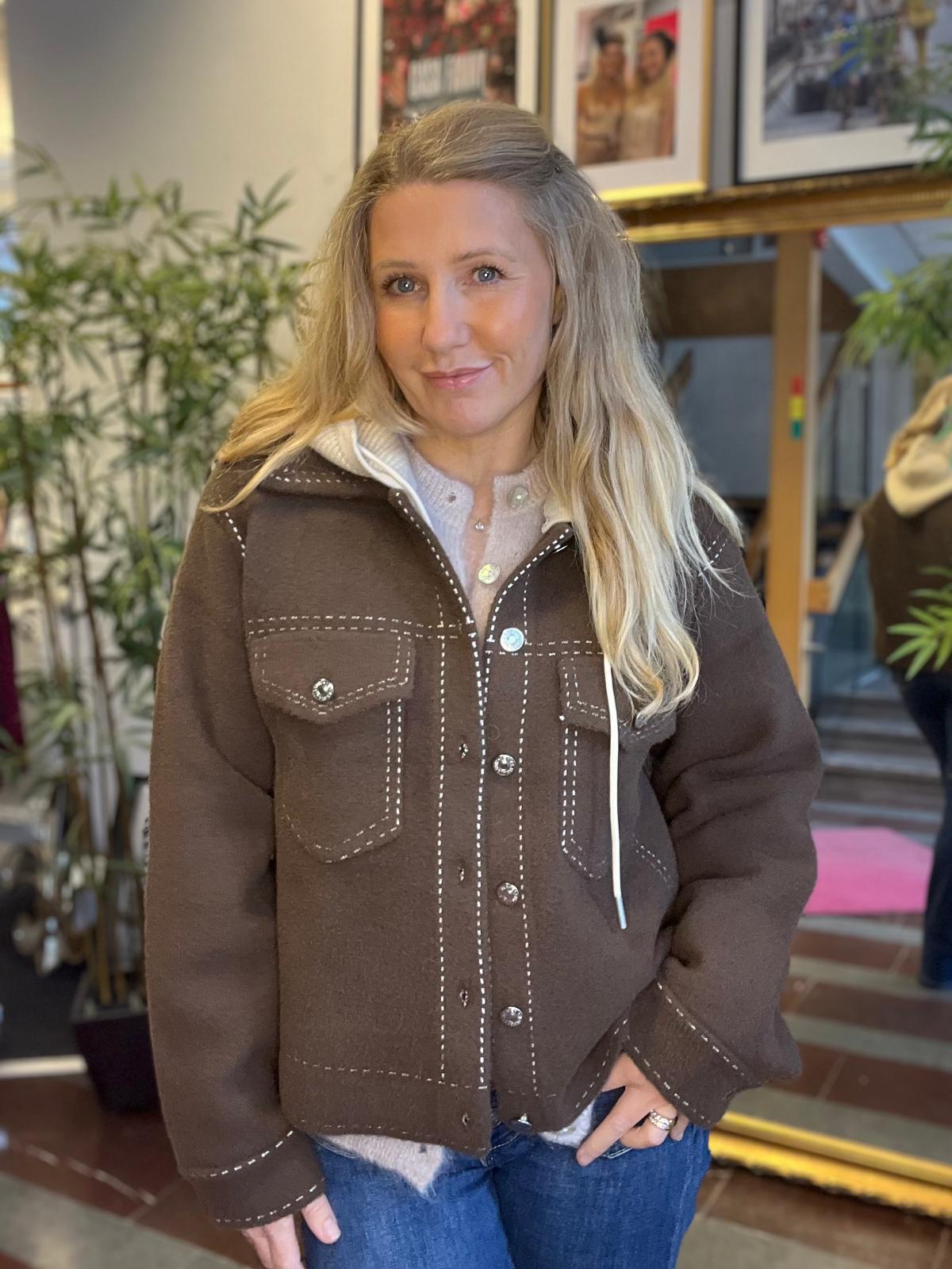 Kvinna med långt blont hår står och poserar inomhus, ser in i kameran och ler svagt. Hon bär brun jacka med vita sömmar och huva samt jeans; bakom syns gröna växter och inramade foton.