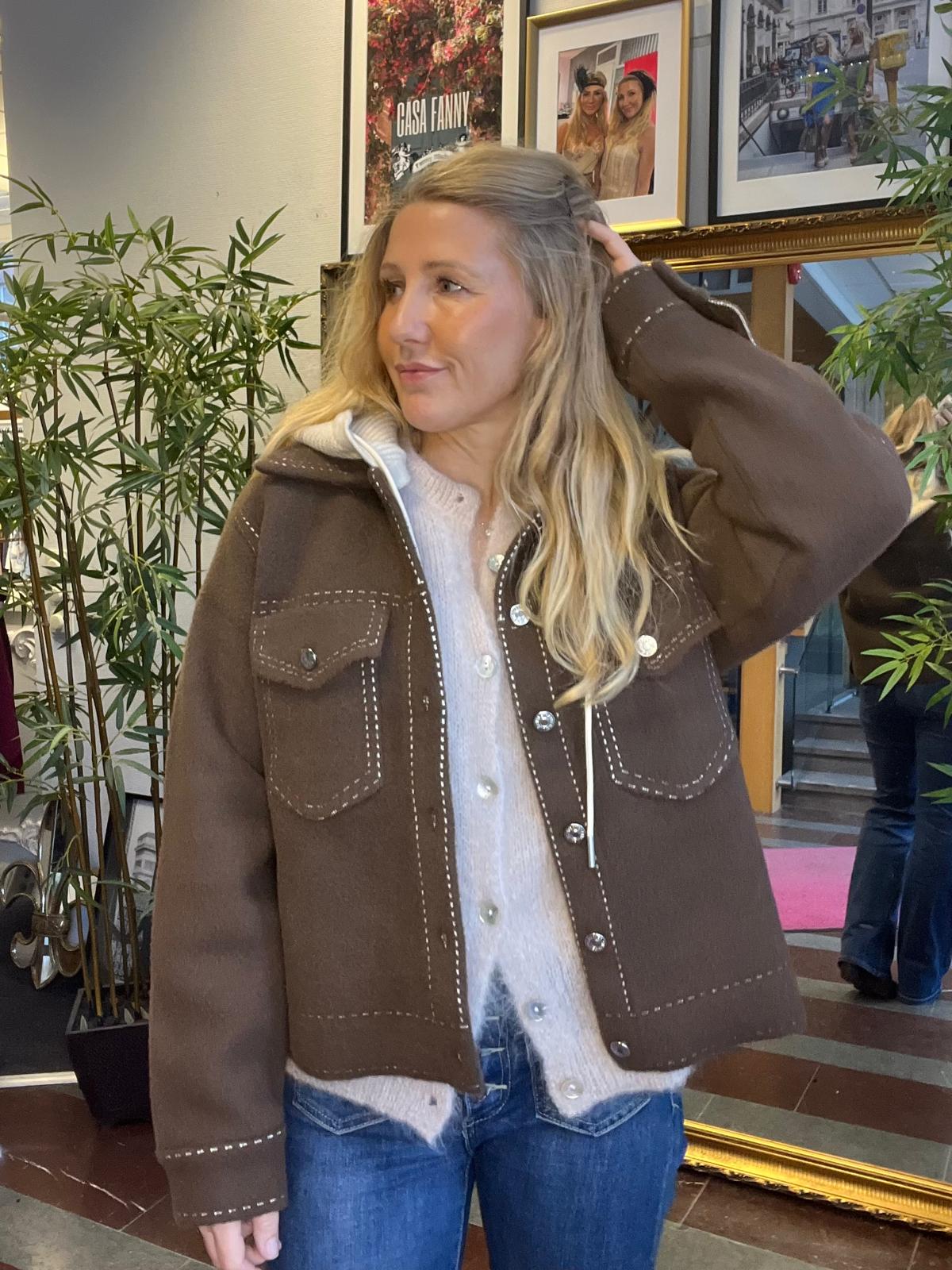 Blond kvinna står inomhus och håller ena handen bakom huvudet medan hon tittar åt sidan. Hon bär brun jacka med vita sömmar över en ljus kofta och jeans, framför gröna växter och inramade foton; texten “CASA FANNY” syns.