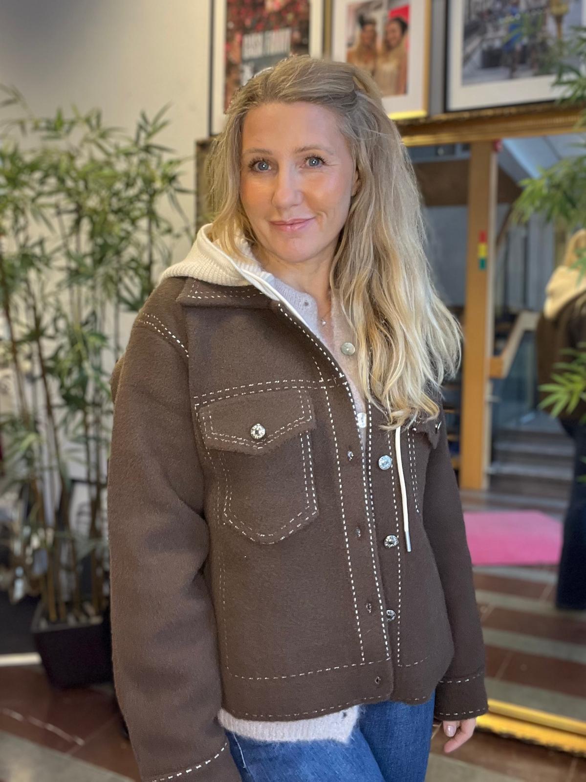 Kvinna med långt blont hår står och ler mot kameran, klädd i brun jacka med vita dekorstickningar över en ljus hoodie och jeans. Inomhusmiljö med gröna krukväxter och inramade foton i bakgrunden.