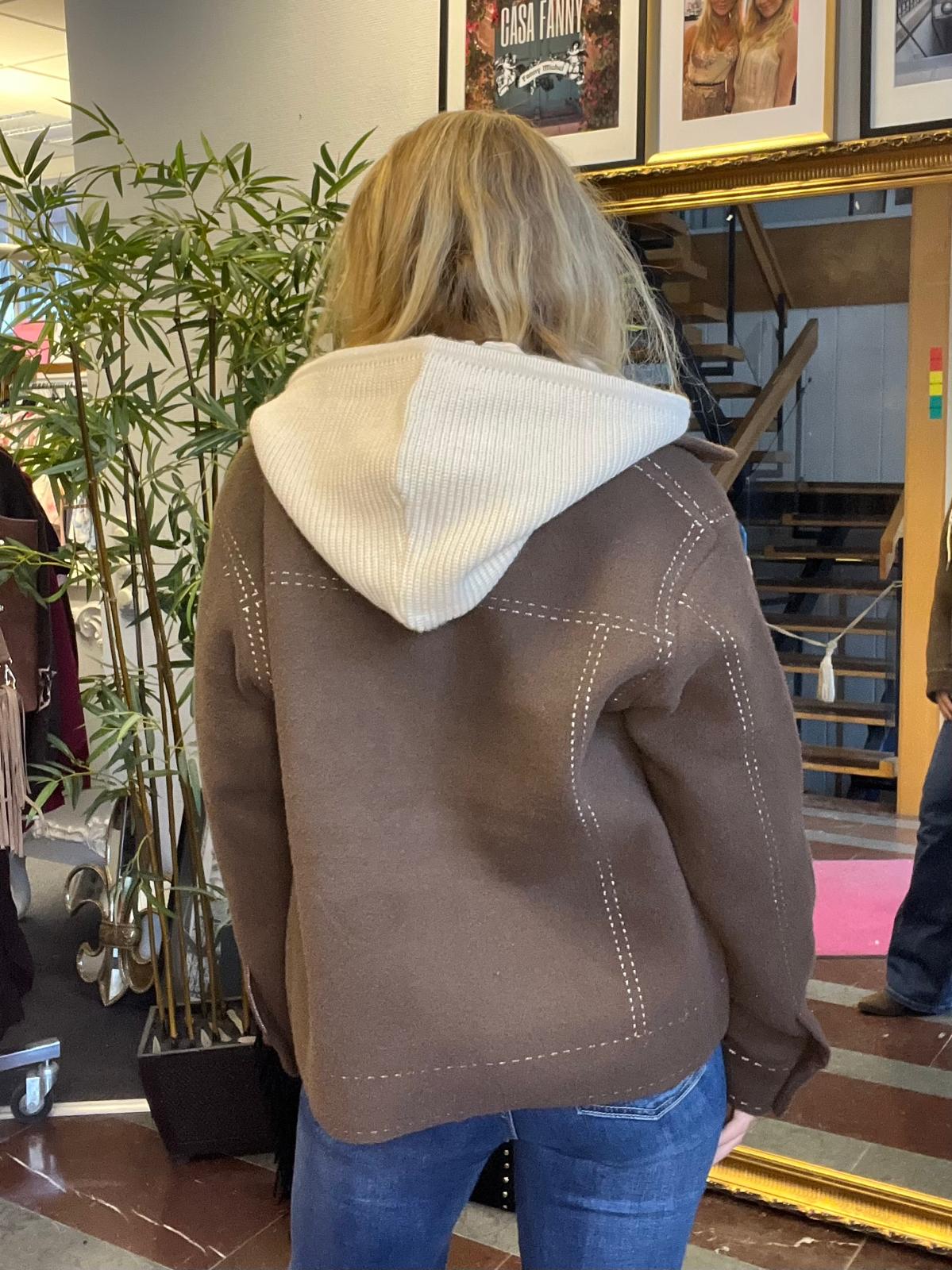 Kvinna med blont hår står med ryggen mot kameran och tittar mot en trappa. Hon bär brun jacka med vita sömmar och vit huva. I bakgrunden syns en stor spegel, bambuväxt och tavlor. Text: “CASA FANNY”.