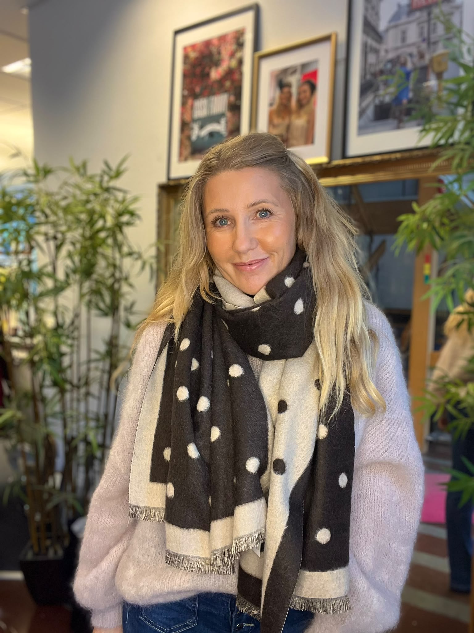 Kvinna med långt blont hår ler mot kameran och står inomhus. Hon bär ljus stickad tröja och en svartvit prickig halsduk. I bakgrunden syns gröna växter och flera inramade fotografier på väggen.