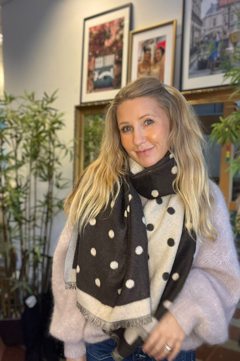 Leende kvinna med långt blont hår står och tittar mot kameran, klädd i ljus stickad tröja och stor svartvit prickig halsduk. I bakgrunden syns gröna krukväxter och inramade foton på väggen.