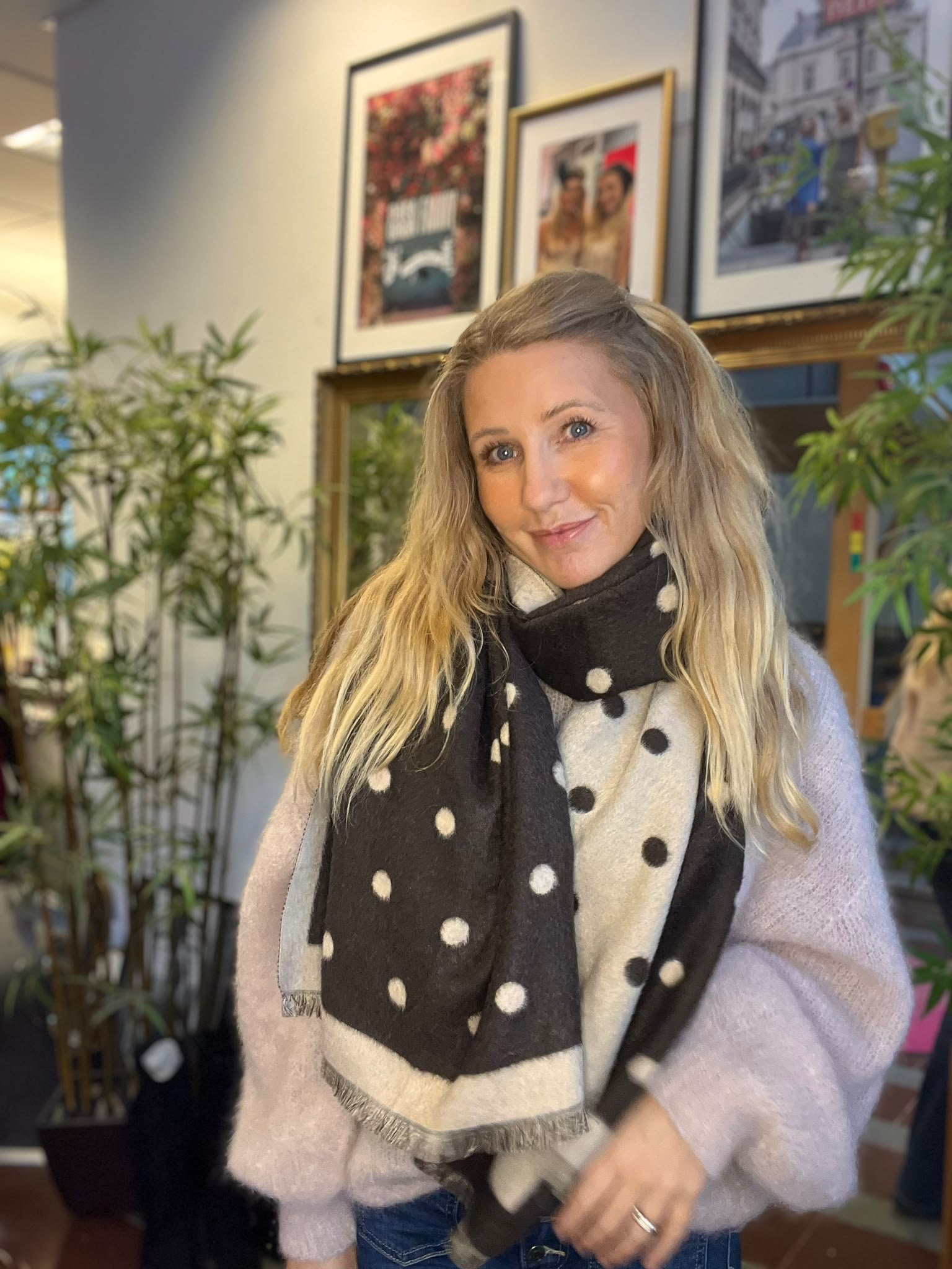 Leende kvinna med långt blont hår står och tittar mot kameran, klädd i ljus stickad tröja och stor svartvit prickig halsduk. I bakgrunden syns gröna krukväxter och inramade foton på väggen.