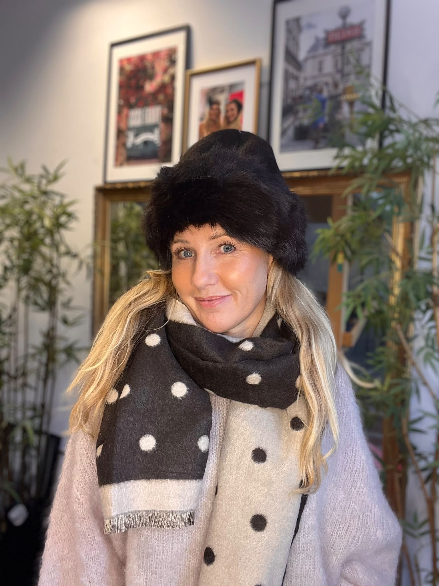 Kvinna i svart pälsmössa och prickig svartvit halsduk ler mot kameran inomhus. Hon står framför en vägg med inramade bilder och gröna krukväxter, i mjukt ljus som ger varm stämning.