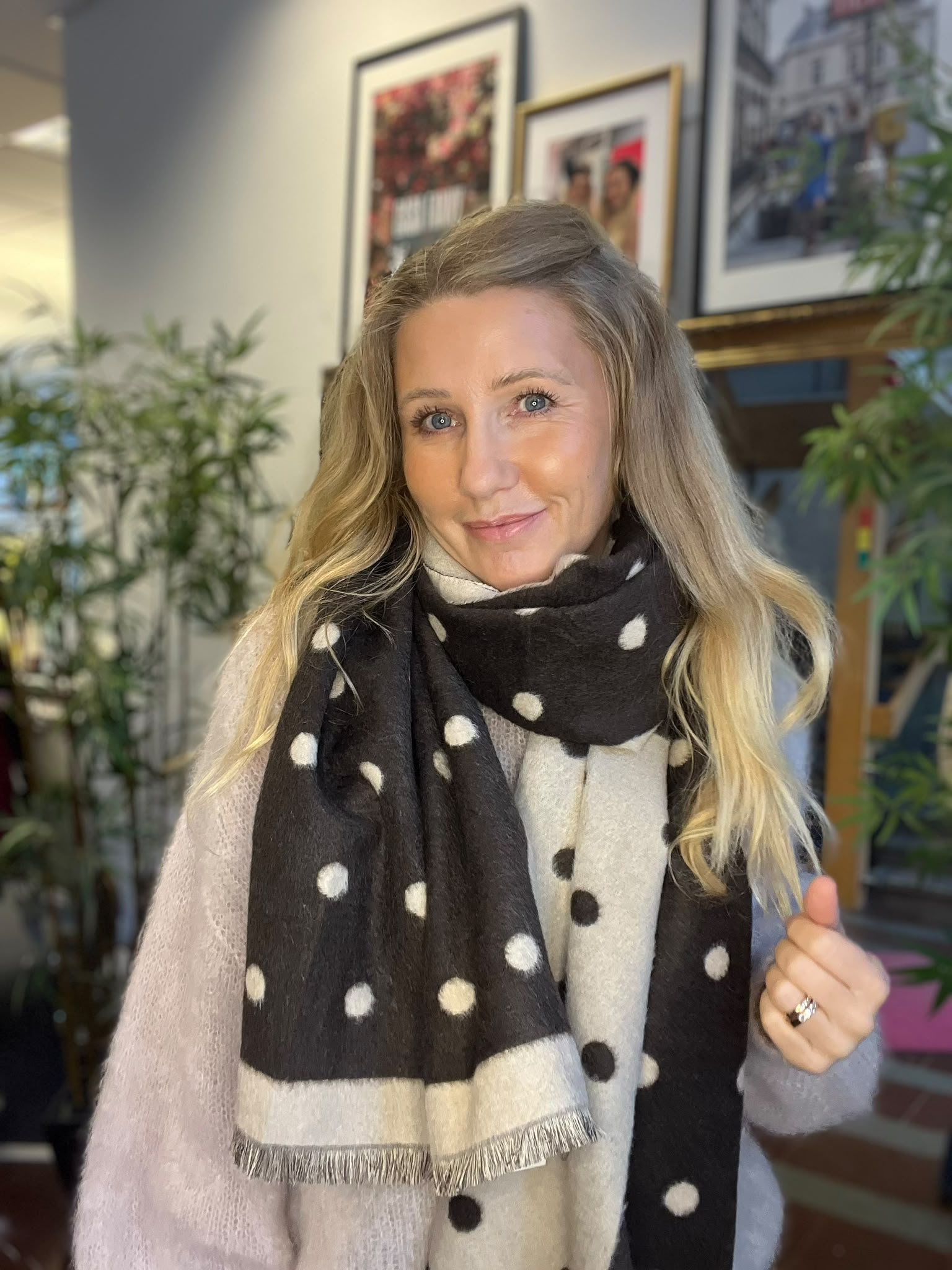 Leende kvinna med långt blont hår tittar mot kameran och håller upp tummen. Hon bär en beige tröja och en stor svartvit prickig scarf, inomhus bland gröna växter och inramade tavlor.