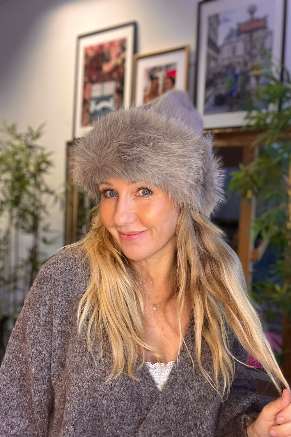 Kvinna med långt blont hår ler mot kameran och bär en grå, fluffig vintermössa samt grå stickad tröja. Hon står inomhus framför inramade affischer och gröna växter i ett varmt belyst rum.