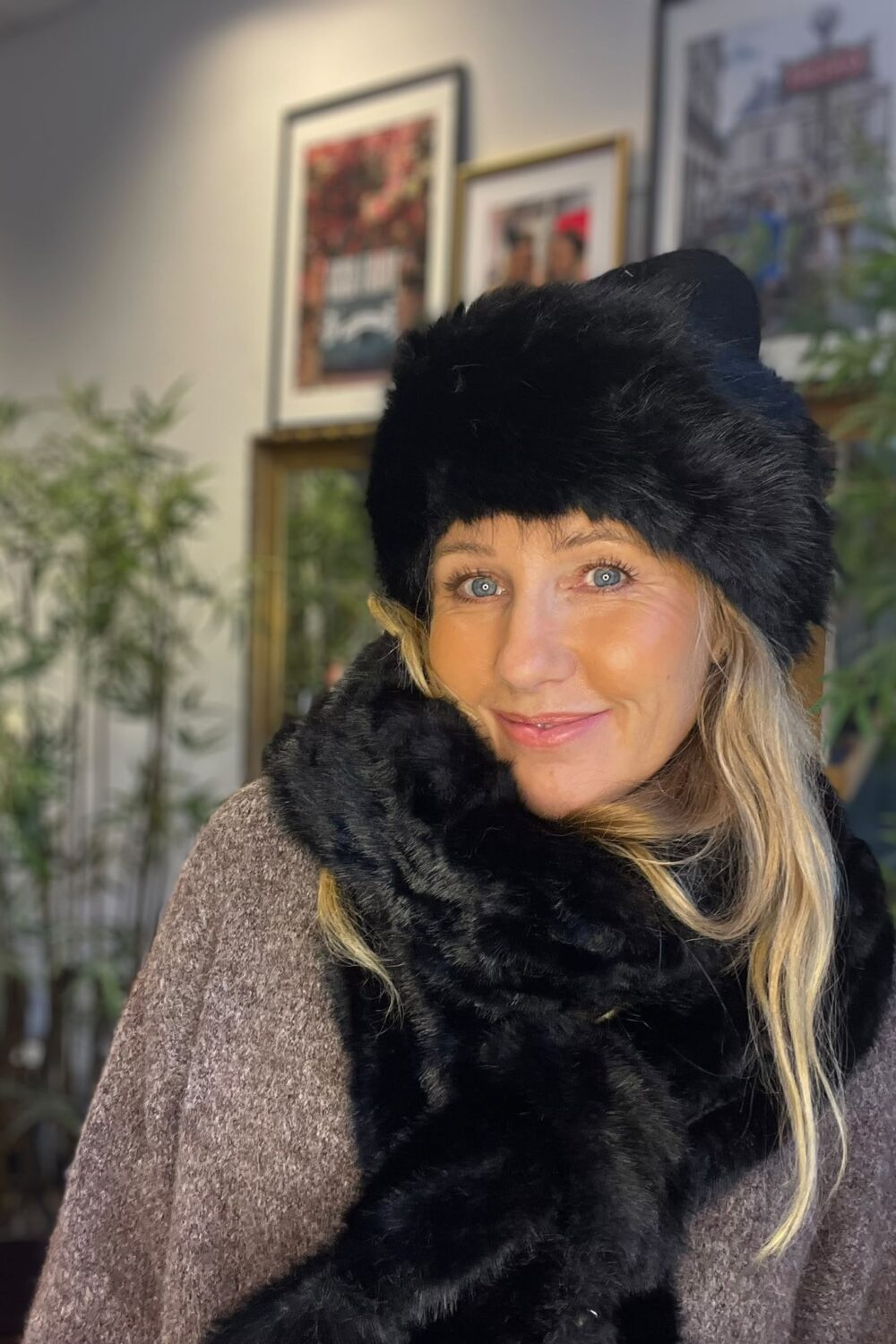 Leende kvinna tittar mot kameran, klädd i svart pälsmössa och tjock, svart pälsig halsduk över en brun stickad tröja. Hon står inomhus framför suddiga växter och inramade tavlor på väggen.