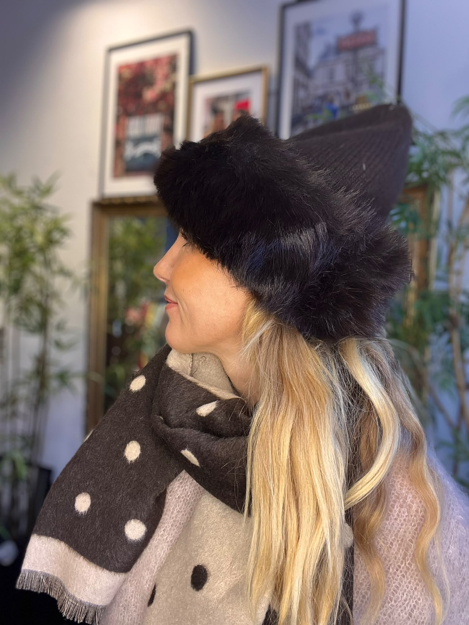 Kvinna i profil bär en stor svart pälsmössa och en prickig halsduk, och tittar åt vänster. Inomhusmiljö med suddiga inramade tavlor och gröna växter i bakgrunden.