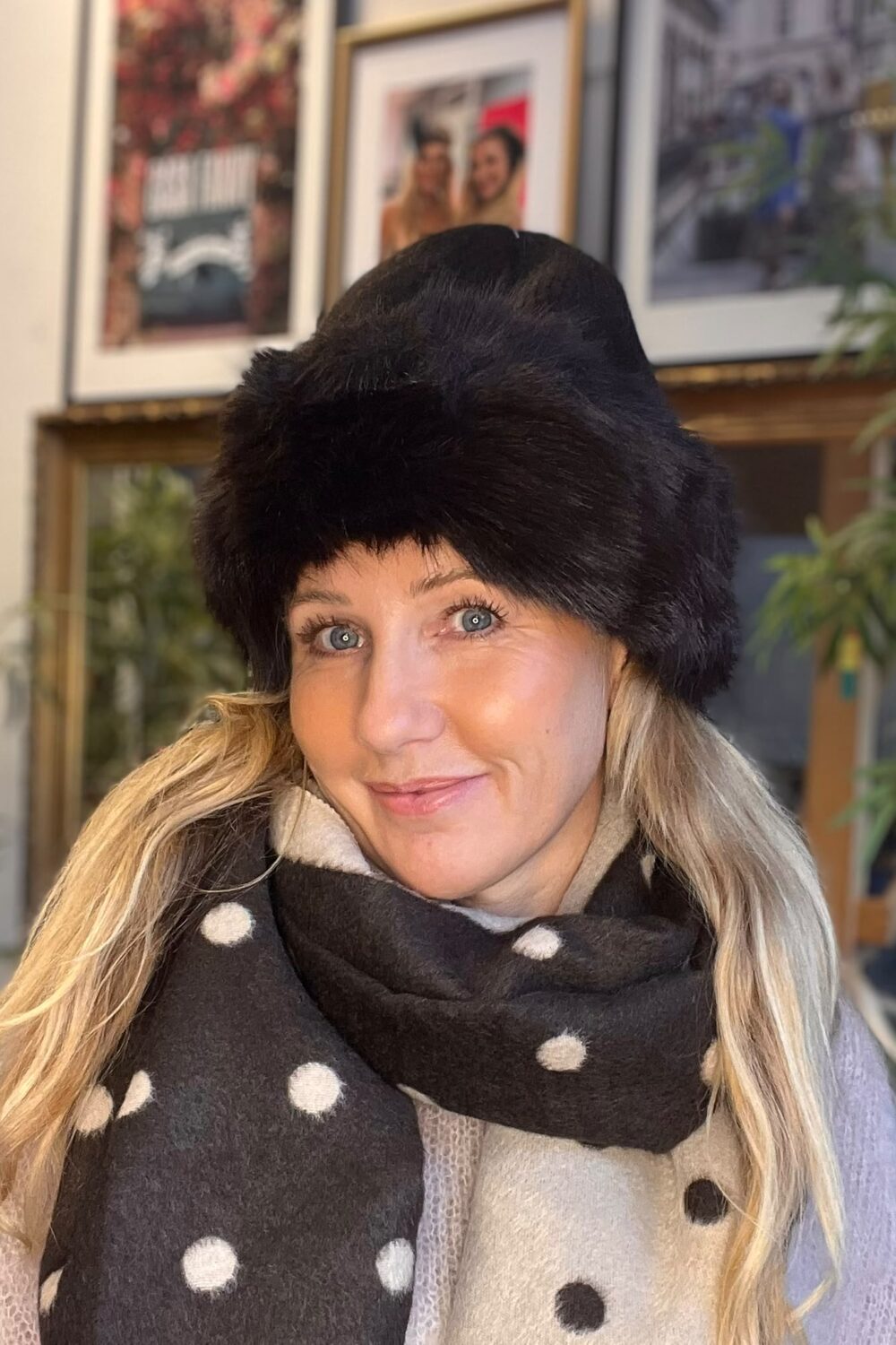 Kvinna i närbild ler mot kameran, klädd i stor svart pälsmössa och tjock prickig halsduk i svart och beige. Inomhus syns inramade foton på väggen och gröna växter i bakgrunden.