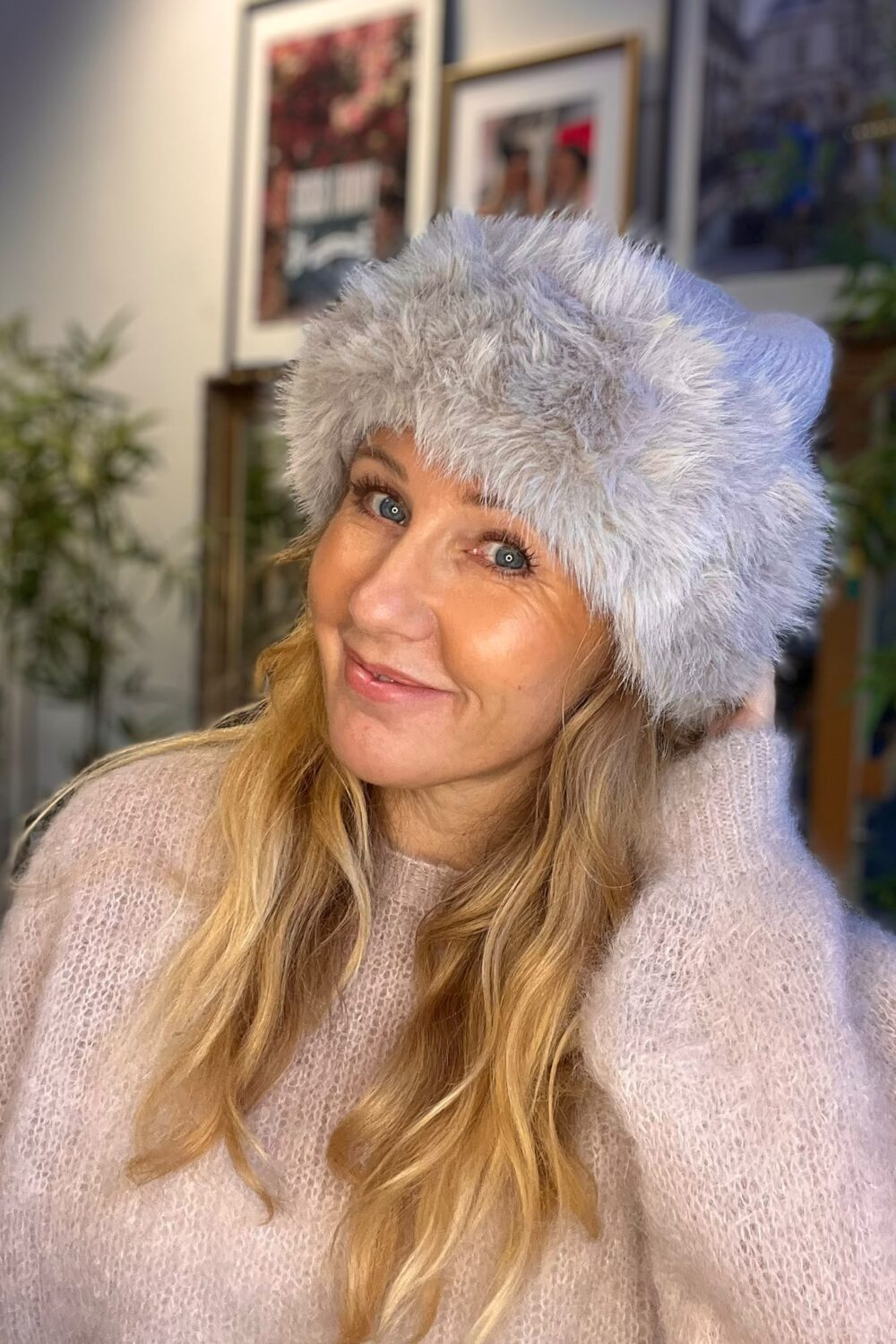 Leende kvinna med långt blont hår poserar med handen bakom huvudet och bär en stor, fluffig grå pälsmössa och ljus stickad tröja. Inomhusmiljö med suddiga tavlor och gröna växter i bakgrunden.