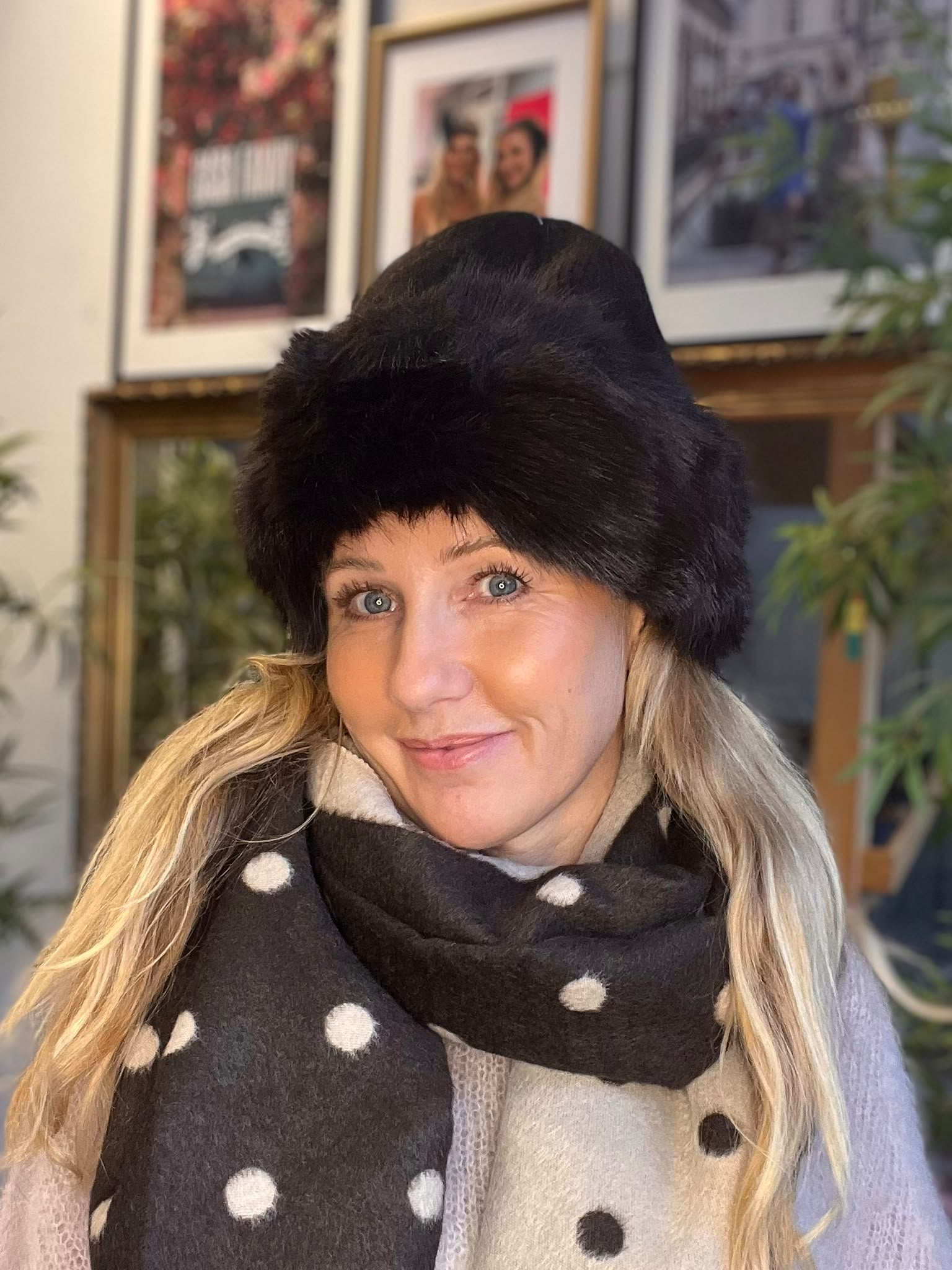 Kvinna i närbild ler mot kameran, klädd i stor svart pälsmössa och tjock prickig halsduk i svart och beige. Inomhus syns inramade foton på väggen och gröna växter i bakgrunden.