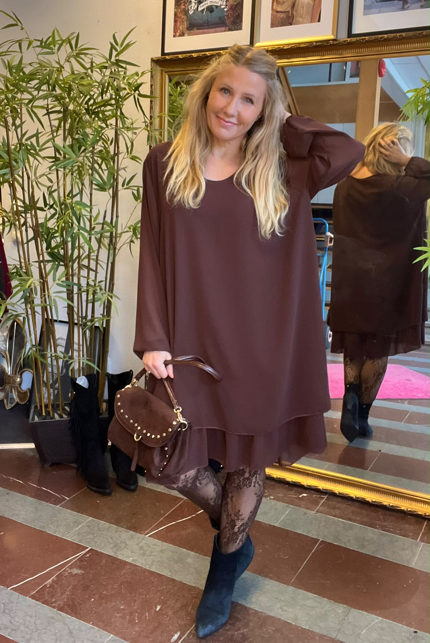 Leende kvinna poserar framför en stor guldramsspegel, med ena handen i håret och den andra som håller en brun handväska. Hon bär en vinröd klänning, spetsstrumpbyxor och svarta boots, bredvid gröna växter.