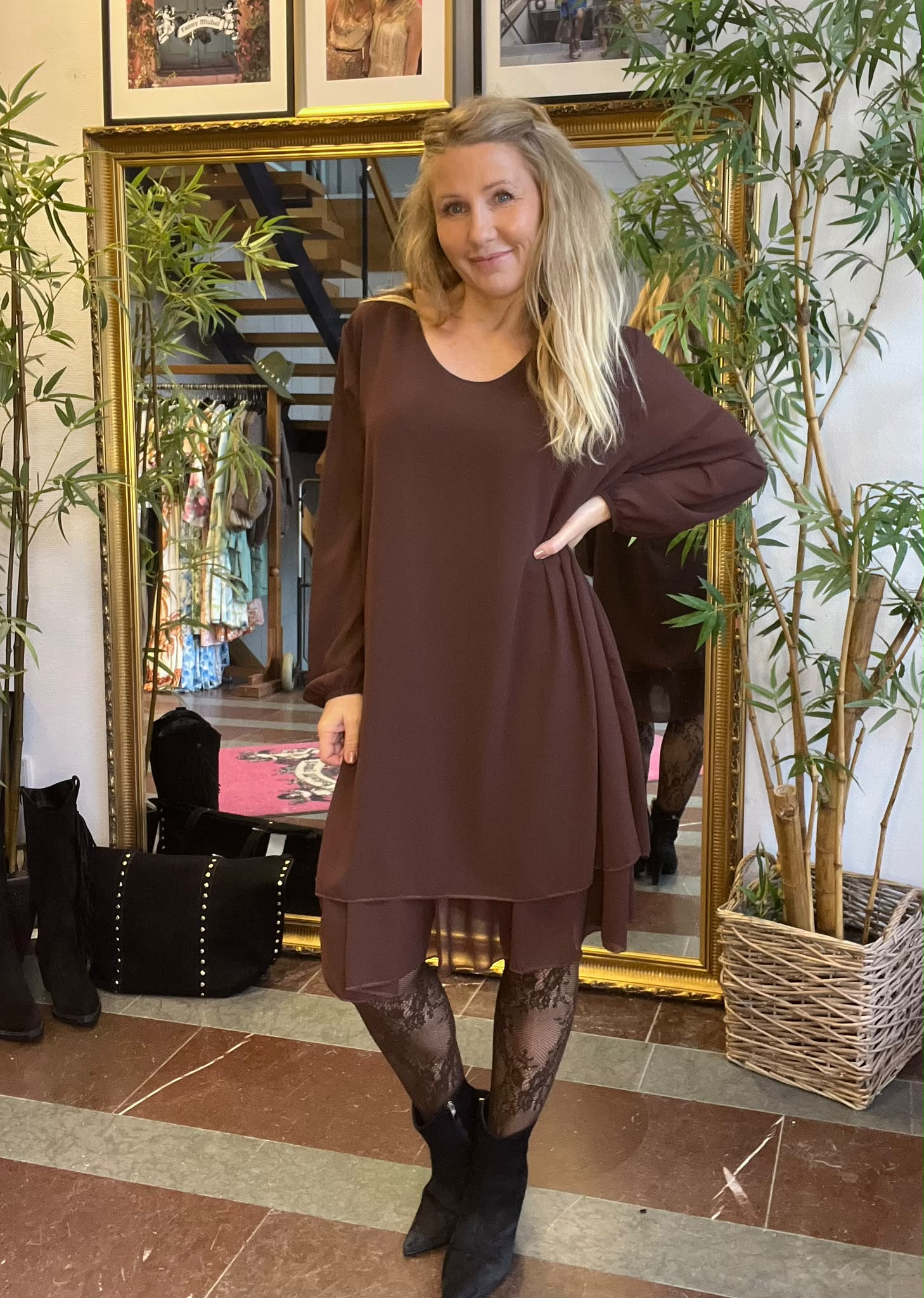 Kvinna står med handen på höften och ler mot kameran, klädd i brun klänning med volang, matchande leggings och spetsiga svarta boots. Hon poserar framför en stor guldramsspegel bland gröna växter.