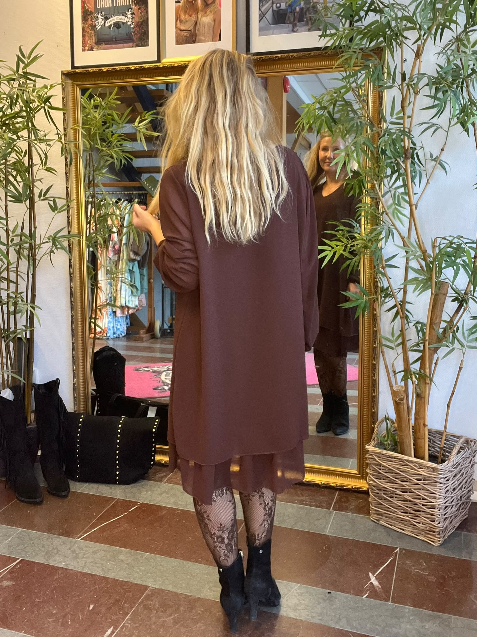 Kvinna med långt blont hår står framför en stor guldramsspegel och tittar på sin spegelbild, som ler. Hon bär en brun klänning, mönstrade strumpbyxor och svarta boots; växter och kläder syns i bakgrunden.