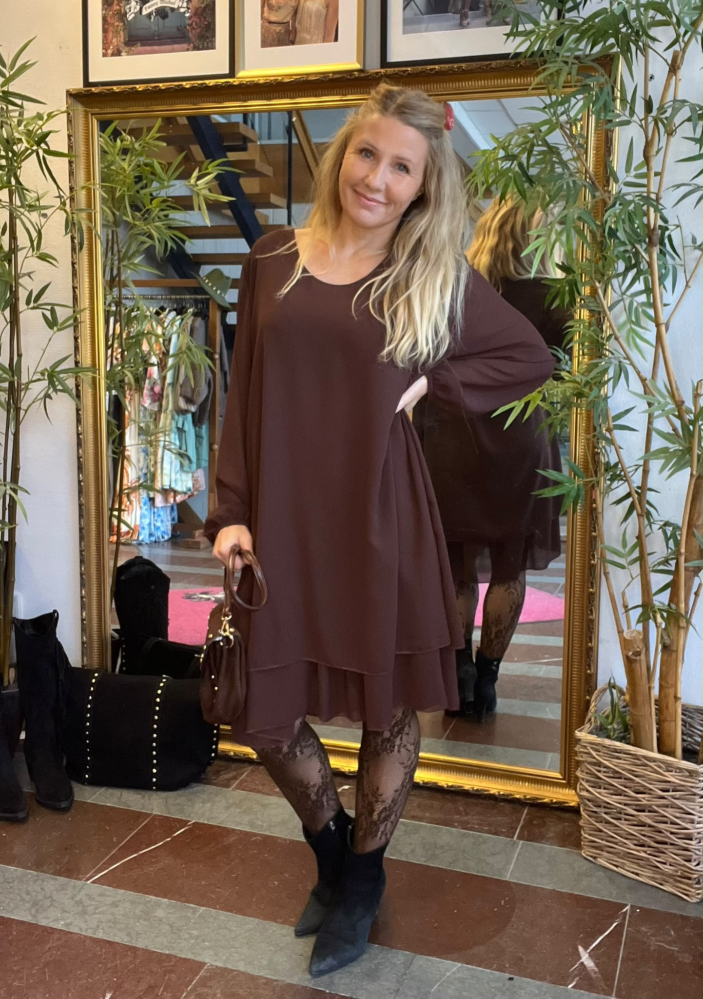 Leende kvinna poserar framför en stor guldramsspegel med handen på höften och en brun handväska i andra handen. Hon bär vinröd tunika, spetsiga svarta stövlar och mönstrade tights, omgiven av gröna växter.