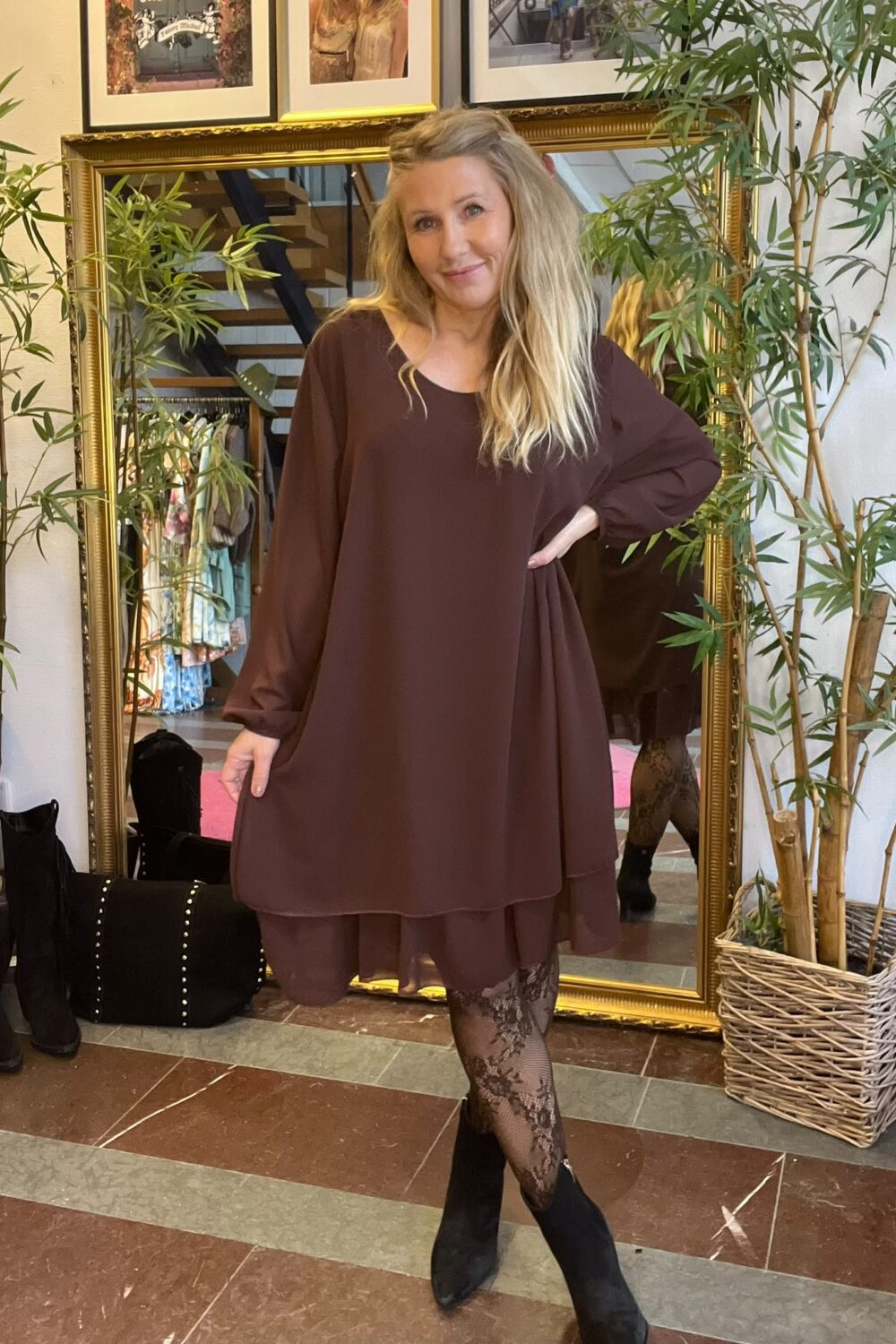 Kvinna med långt blont hår poserar med handen på höften i en brun, lös klänning, mönstrade strumpbyxor och svarta boots. Hon står framför en stor guldramsspegel, omgiven av gröna växter och klädställning.