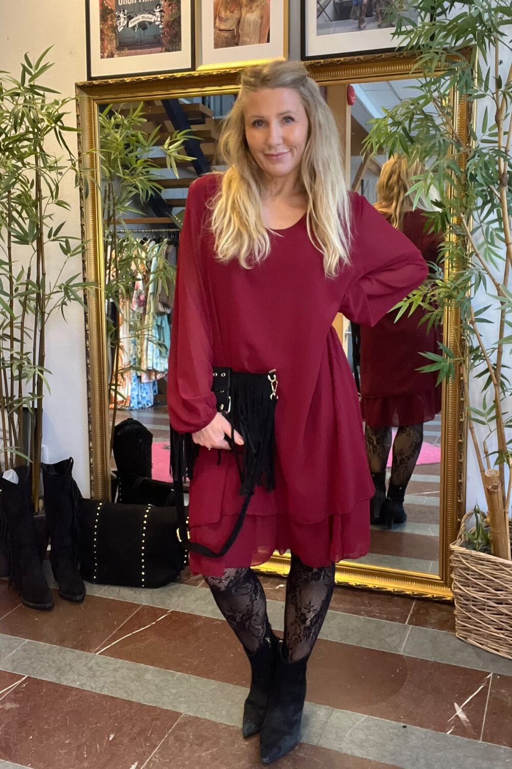 Kvinna med långt blont hår poserar med handen på höften framför en stor guldramsspegel, klädd i vinröd klänning, svarta spetsstrumpbyxor och boots. Hon håller en svart fransväska; bambuplantor och skor syns i butiken. Text: “CASA FANNY”.