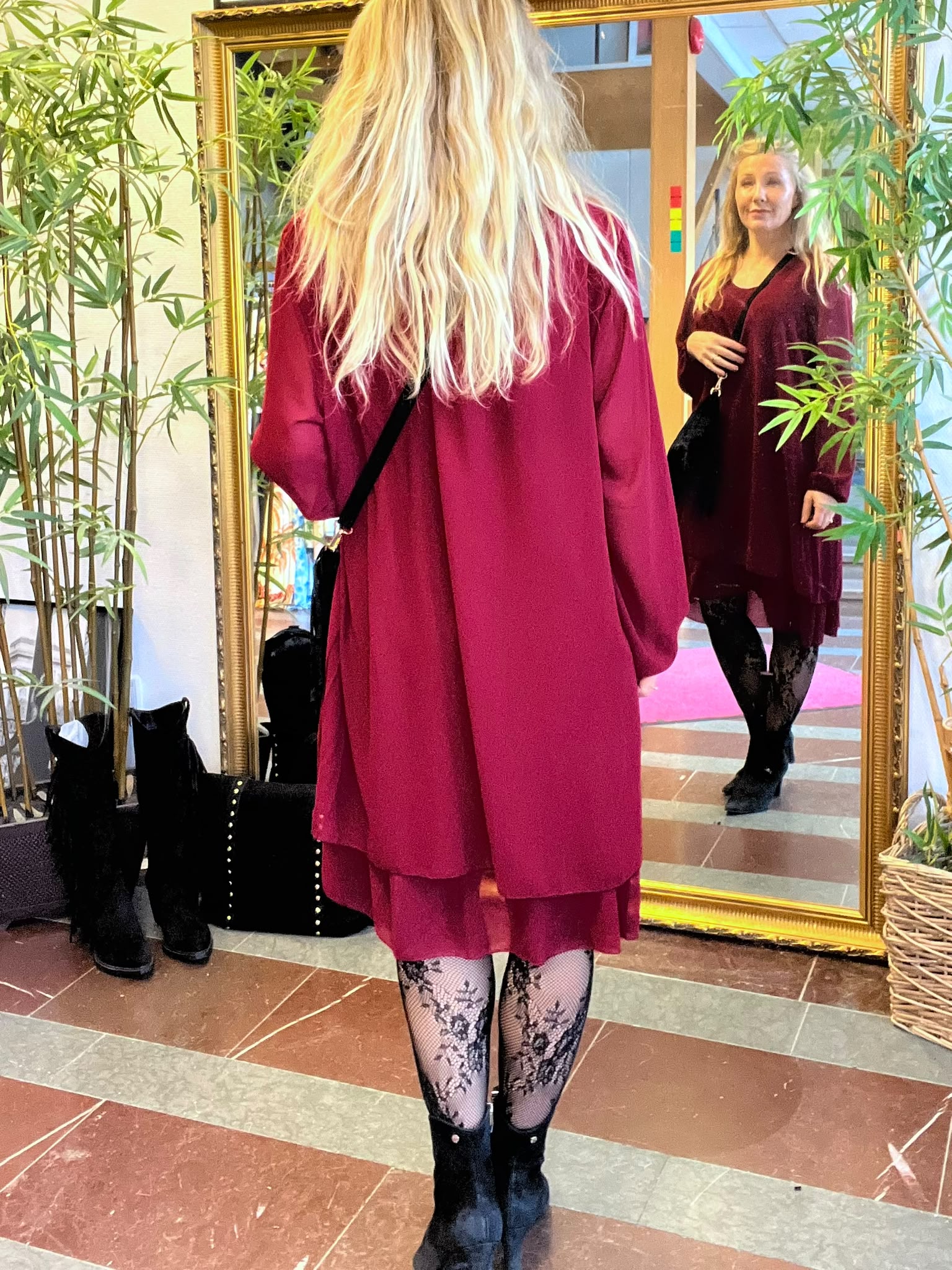 Kvinna med långt blont hår står framför en stor guldramad spegel och ser sin spegelbild. Hon bär vinröd klänning, svarta stövlar och mönstrade strumpbyxor; växter och en väska syns bredvid.