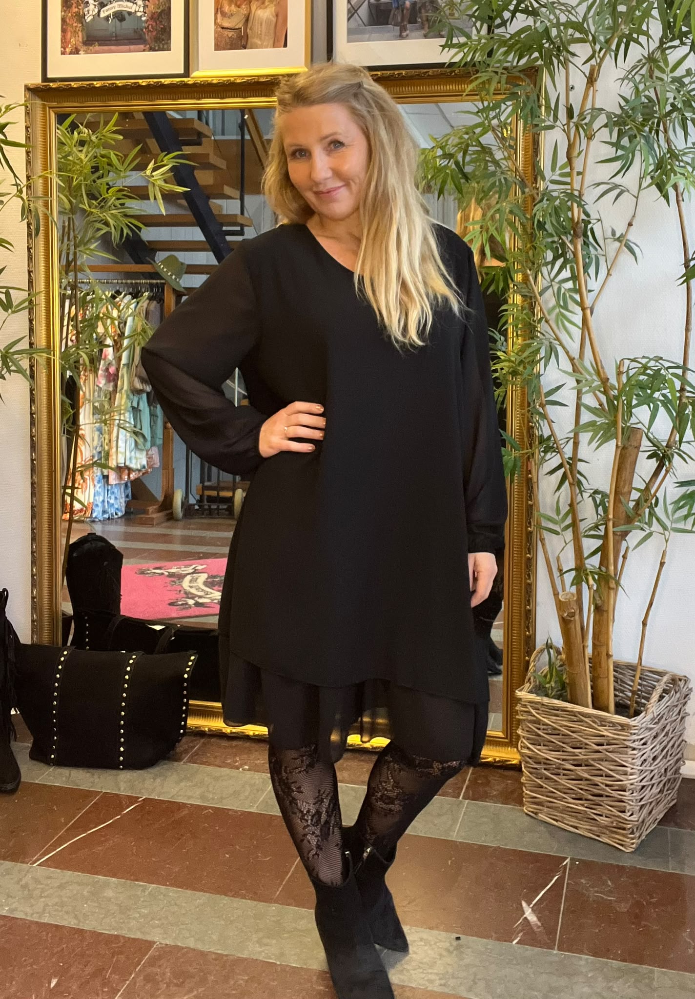 Blond kvinna i svart långärmad klänning poserar med handen på höften och ler mot kameran. Hon står framför en stor guldramsspegel, med gröna krukväxter och en klädbutiksmiljö i bakgrunden.