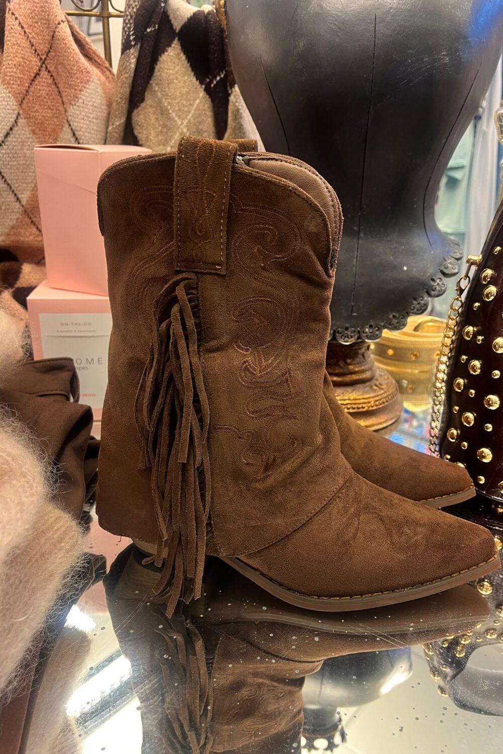 Brun mockastövel i cowboymodell står uppställd på ett spegelbord, med broderade mönster och långa fransar på sidan. I bakgrunden syns en svart skyltdocka, rutiga sjalar och en nitarbetad väska.