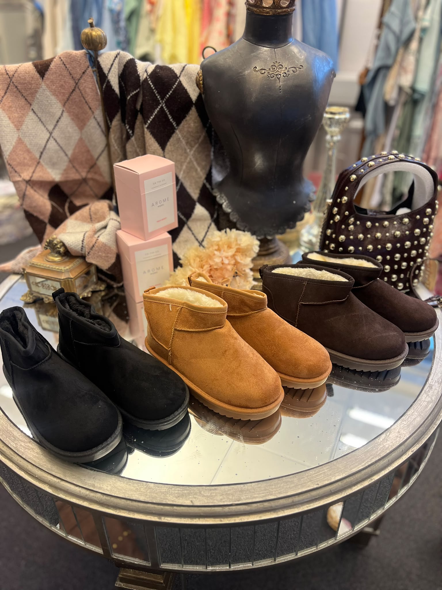 Butiksdisplay på runt spegelbord visar fodrade ankelboots i svart, cognac och mörkbrunt, arrangerade framför en svart provdocka. Bakom ligger rutiga sjalar, en nitarprydd väska och rosa askar med texten “AROME”.