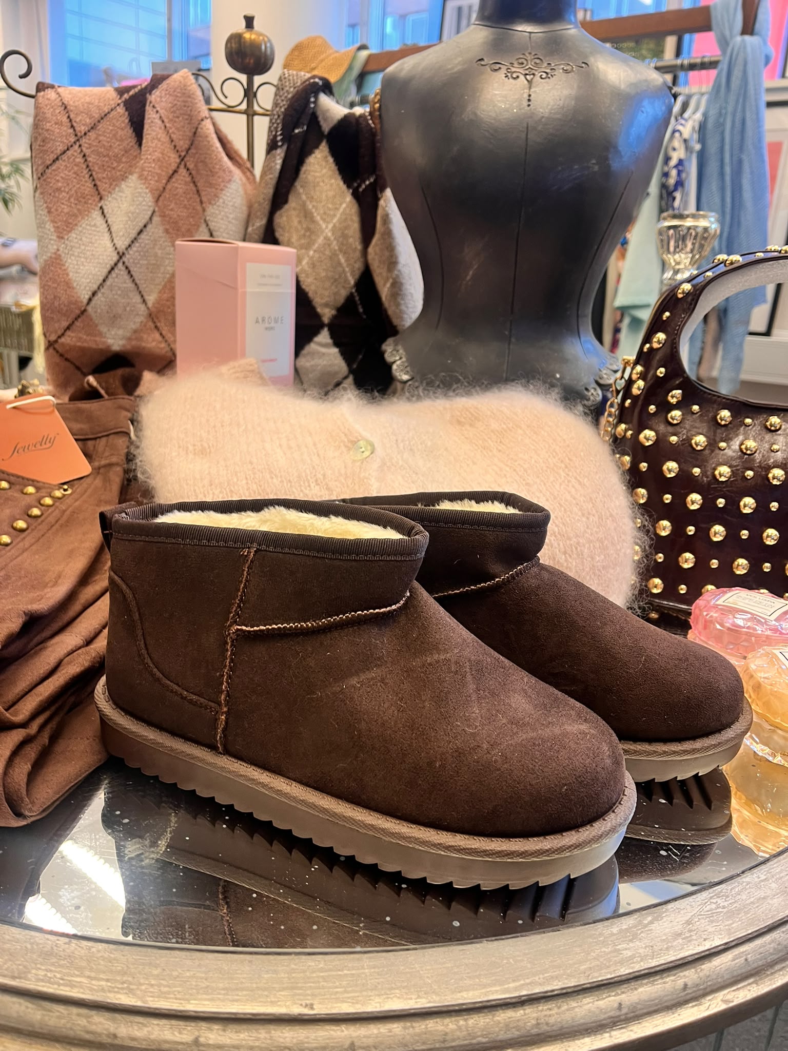 Bruna mockaboots med ljus teddyfoder står på ett runt spegelbord i en butiksexponering, omgivna av argylemönstrade stickade plagg, en svart skyltdockstorso, nitad brun väska och en rosa ask med texten “ARÔME”.