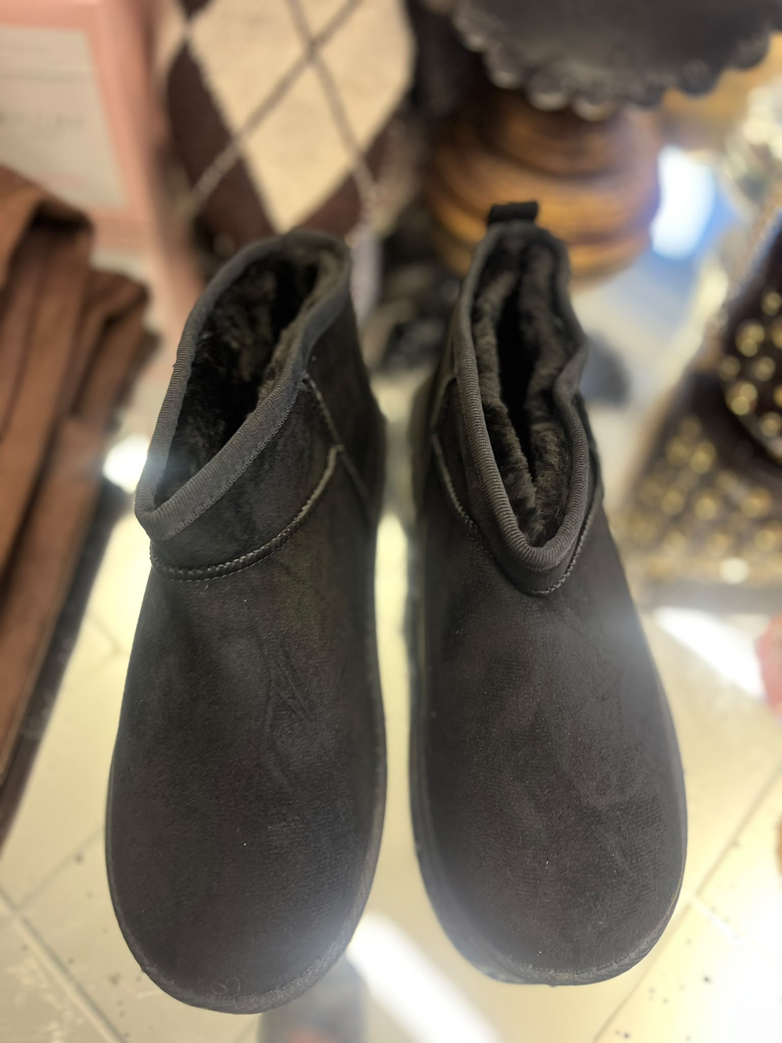 Ett par svarta, fodrade ankelboots står sida vid sida och fotograferas uppifrån på ett ljust golv. Skorna har mjuk, mörk insida och rund tå; i bakgrunden syns suddiga textilier och föremål.