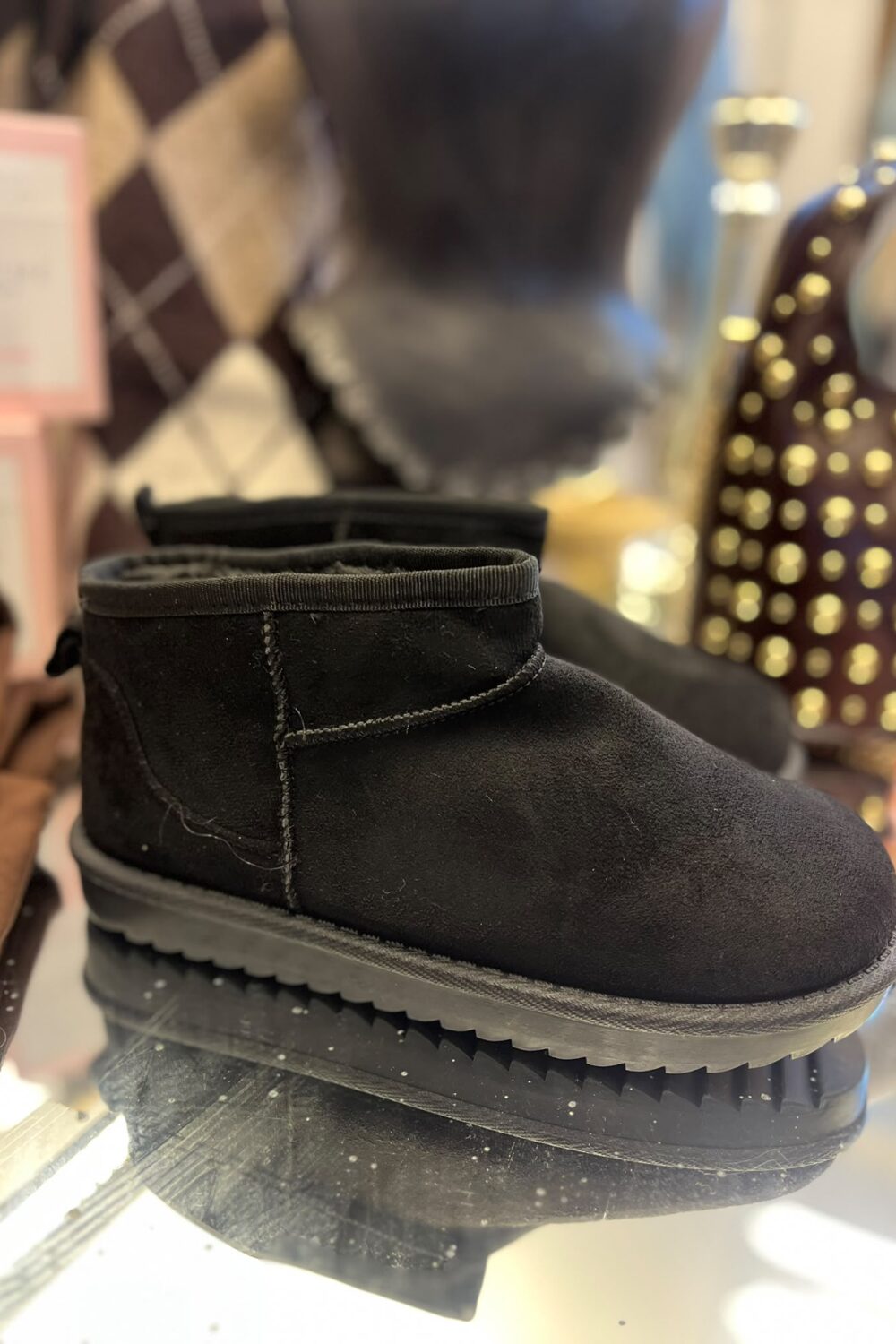 Svart låg ankelstövel i mockaliknande material står på en speglande hylla, med synliga sömmar och grov räfflad sula. I bakgrunden syns suddiga förpackningar och en brun accessoar med guldfärgade nitar.