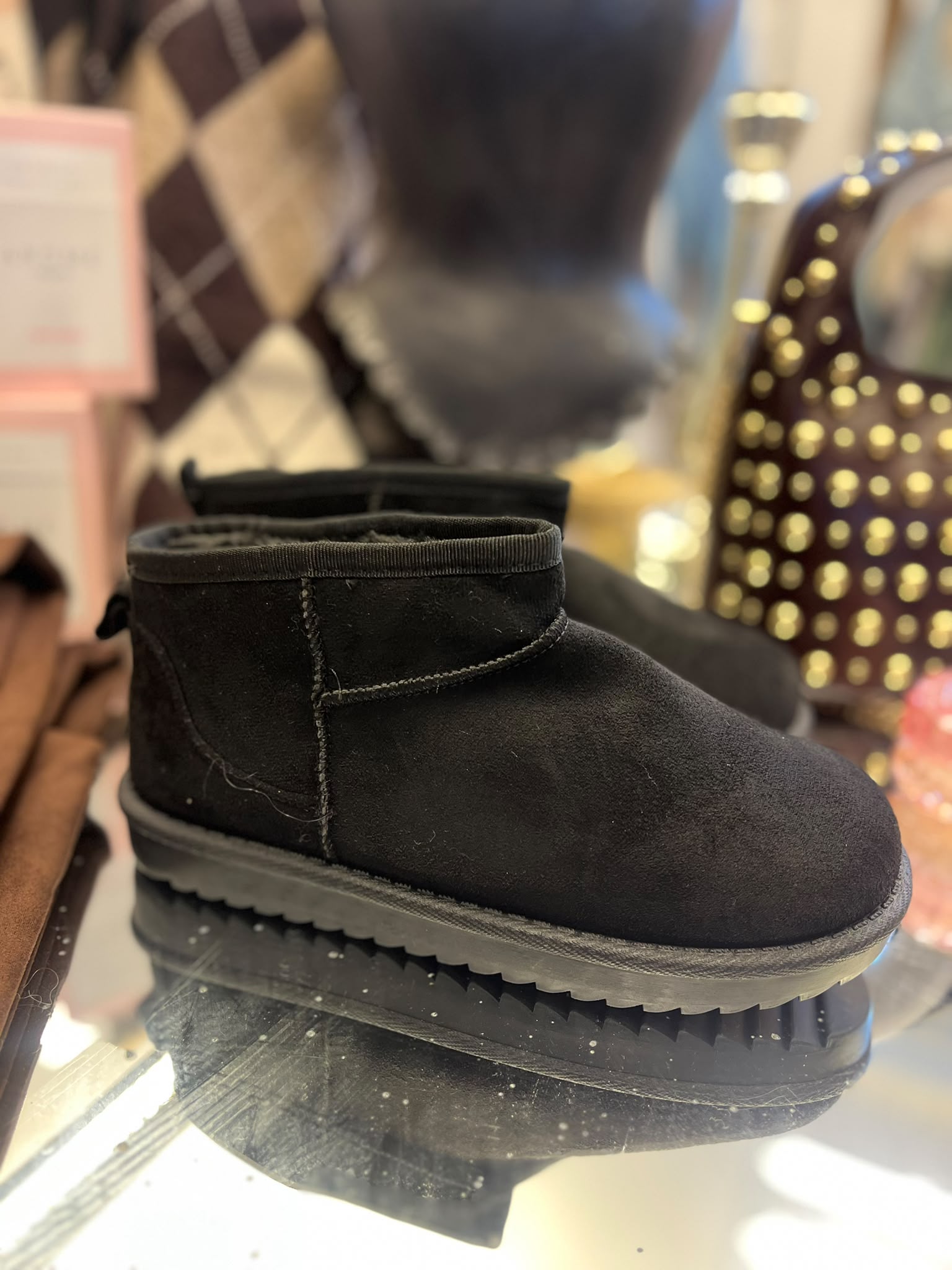 Svart låg ankelstövel i mockaliknande material står på en speglande hylla, med synliga sömmar och grov räfflad sula. I bakgrunden syns suddiga förpackningar och en brun accessoar med guldfärgade nitar.