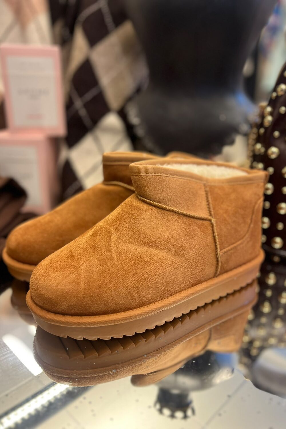 Ett par bruna mockaboots med fluffigt, ljust foder står uppställda på en hylla och visas i närbild. I bakgrunden syns en suddig butiksmiljö med väskor och skyltar.
