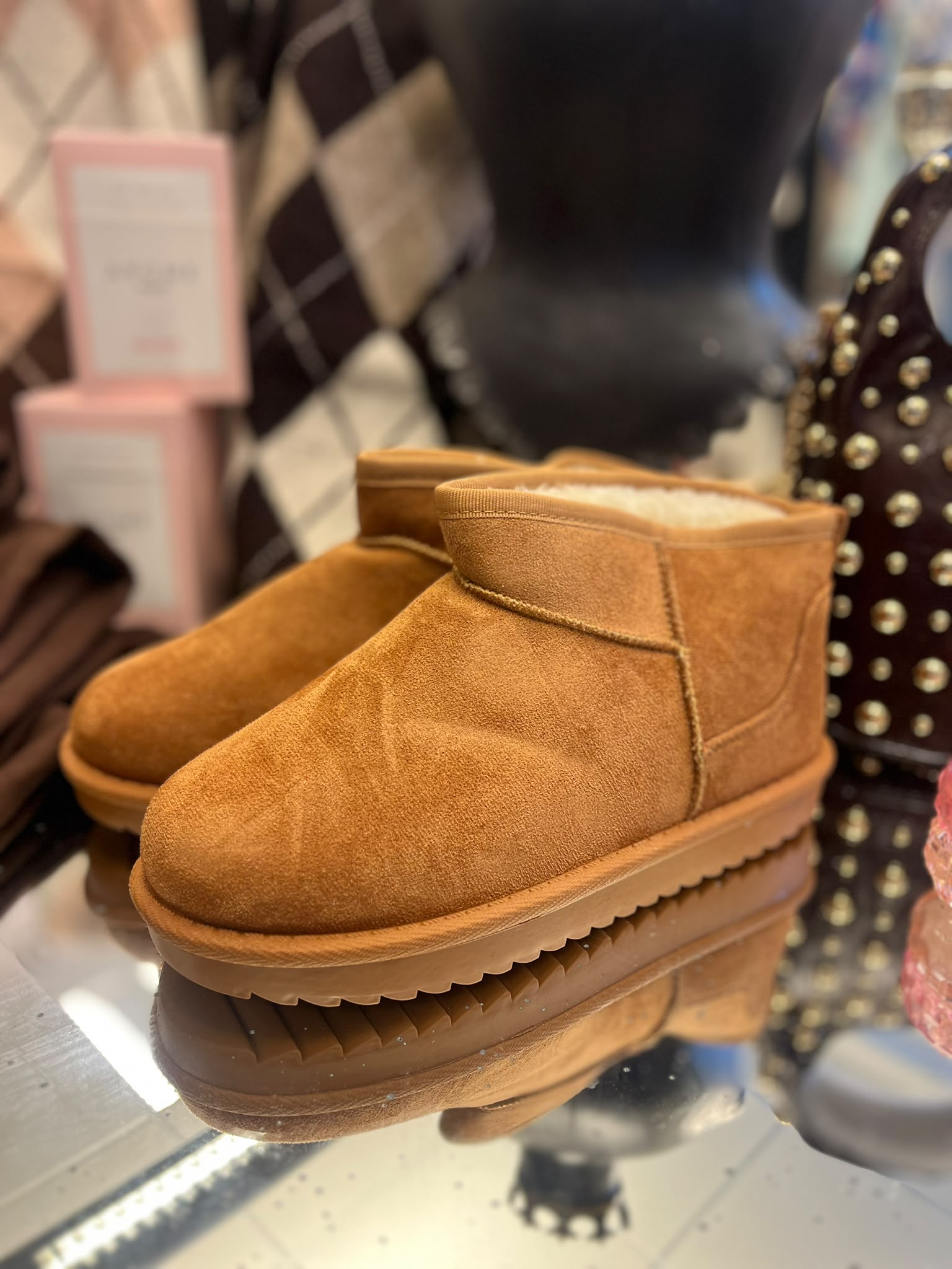 Ett par bruna mockaboots med fluffigt, ljust foder står uppställda på en hylla och visas i närbild. I bakgrunden syns en suddig butiksmiljö med väskor och skyltar.