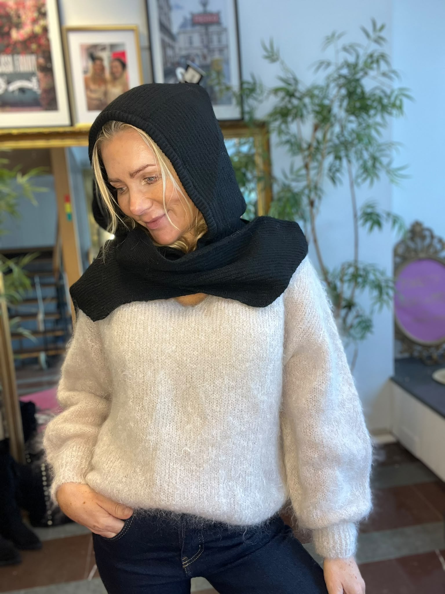 Kvinna med svart stickad huva och halskrage tittar ned och ler lätt, klädd i vit fluffig tröja och jeans. Hon står inomhus framför inramade foton och en grön växt, med varm, hemtrevlig känsla.