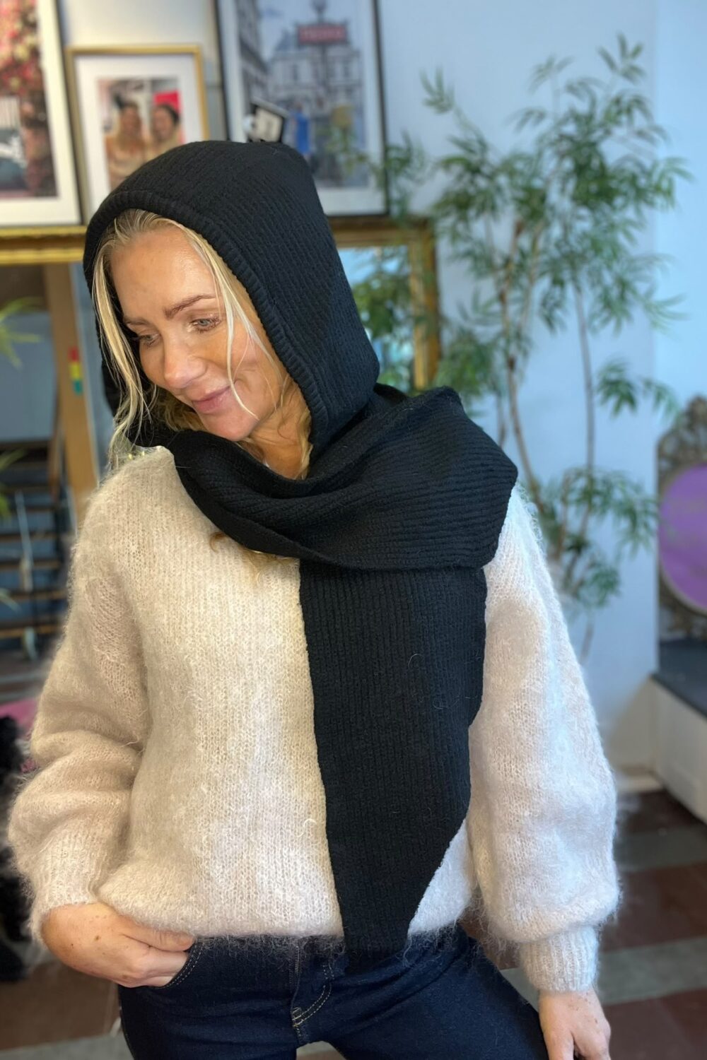 Blond kvinna sitter avslappnat och tittar ned med ett svagt leende, iklädd beige stickad tröja och svart huva med lång halsduk. Inomhusmiljö med inramade tavlor på väggen och en grön växt i bakgrunden.
