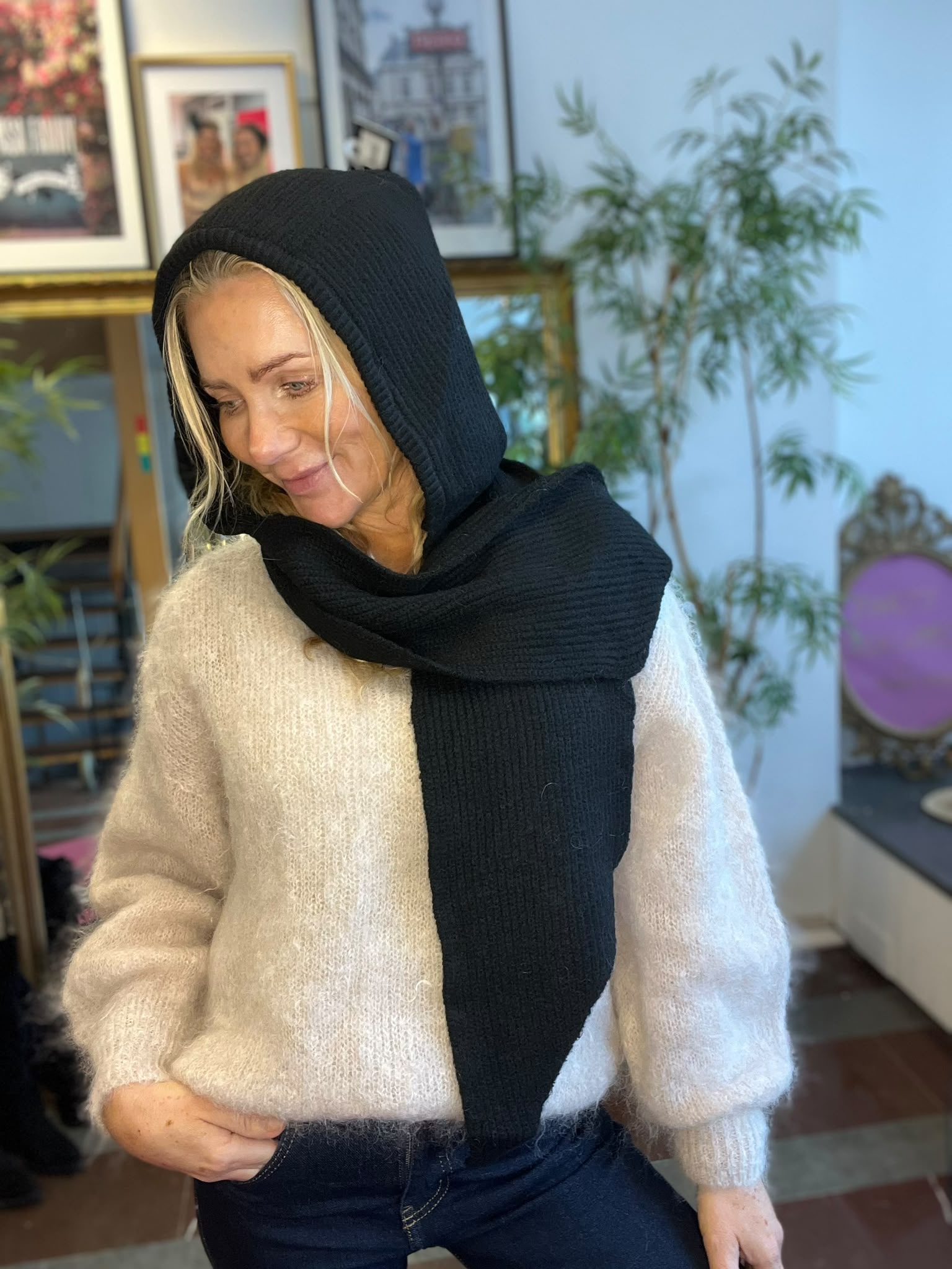 Blond kvinna sitter avslappnat och tittar ned med ett svagt leende, iklädd beige stickad tröja och svart huva med lång halsduk. Inomhusmiljö med inramade tavlor på väggen och en grön växt i bakgrunden.