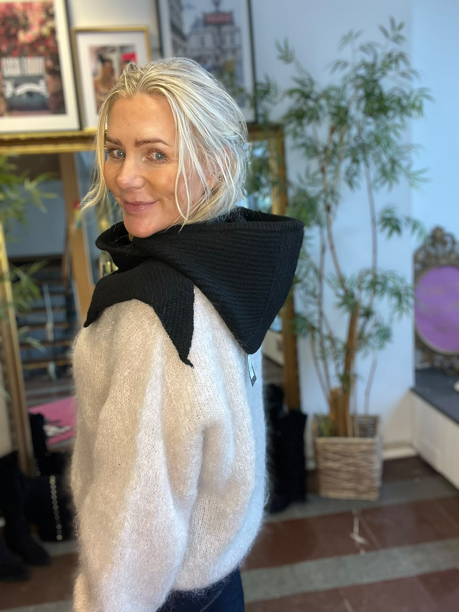 Blond kvinna vänder sig om och ler mot kameran, klädd i ljus stickad tröja med svart sjal över axlarna. Hon står inomhus framför inramade tavlor och en hög, grön krukväxt.