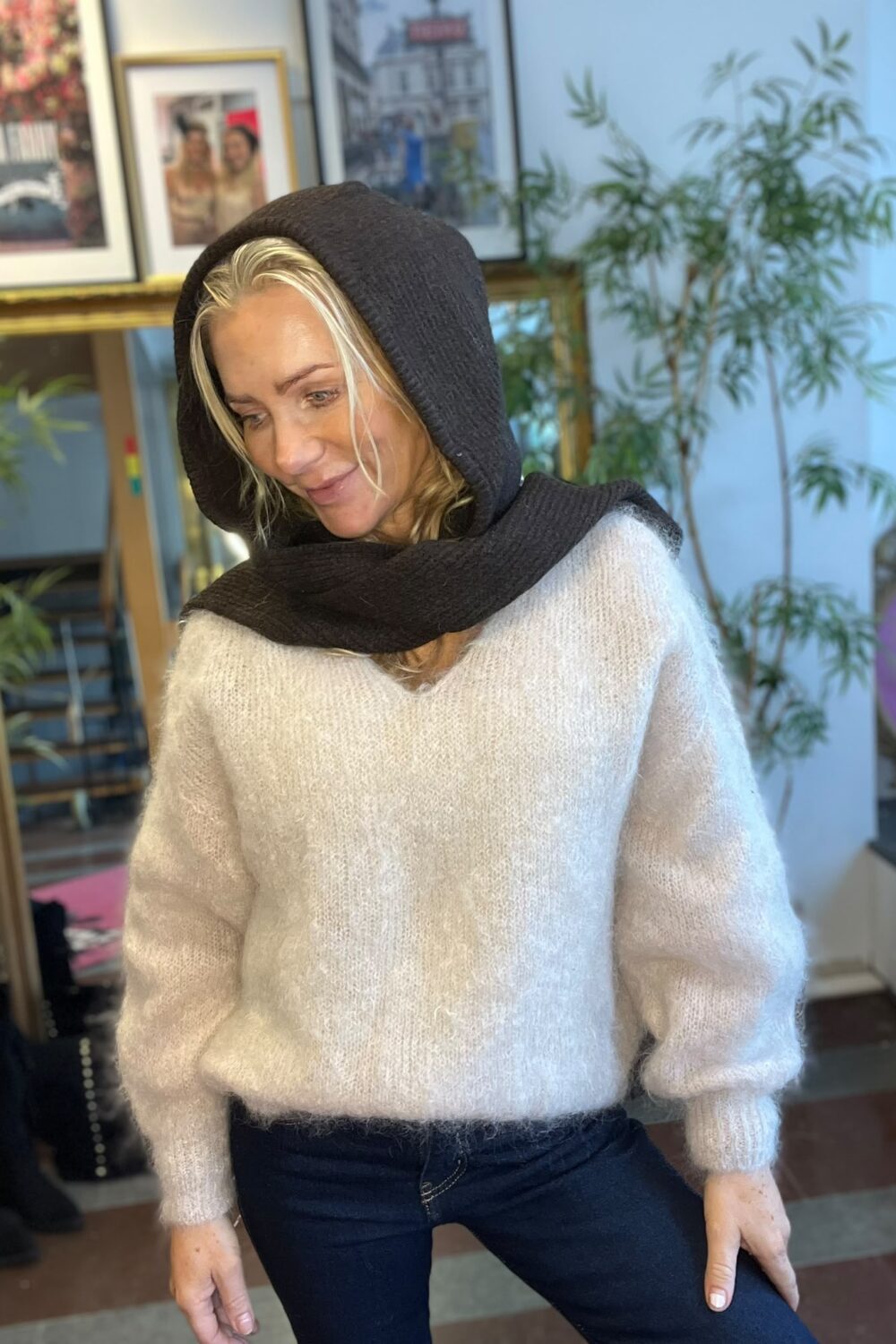 Blond kvinna lutar sig lätt fram och tittar ned med ett svagt leende, iklädd ljus stickad tröja och mörk huvsjal. Inomhusmiljö med inramade affischer och en grön växt i bakgrunden.