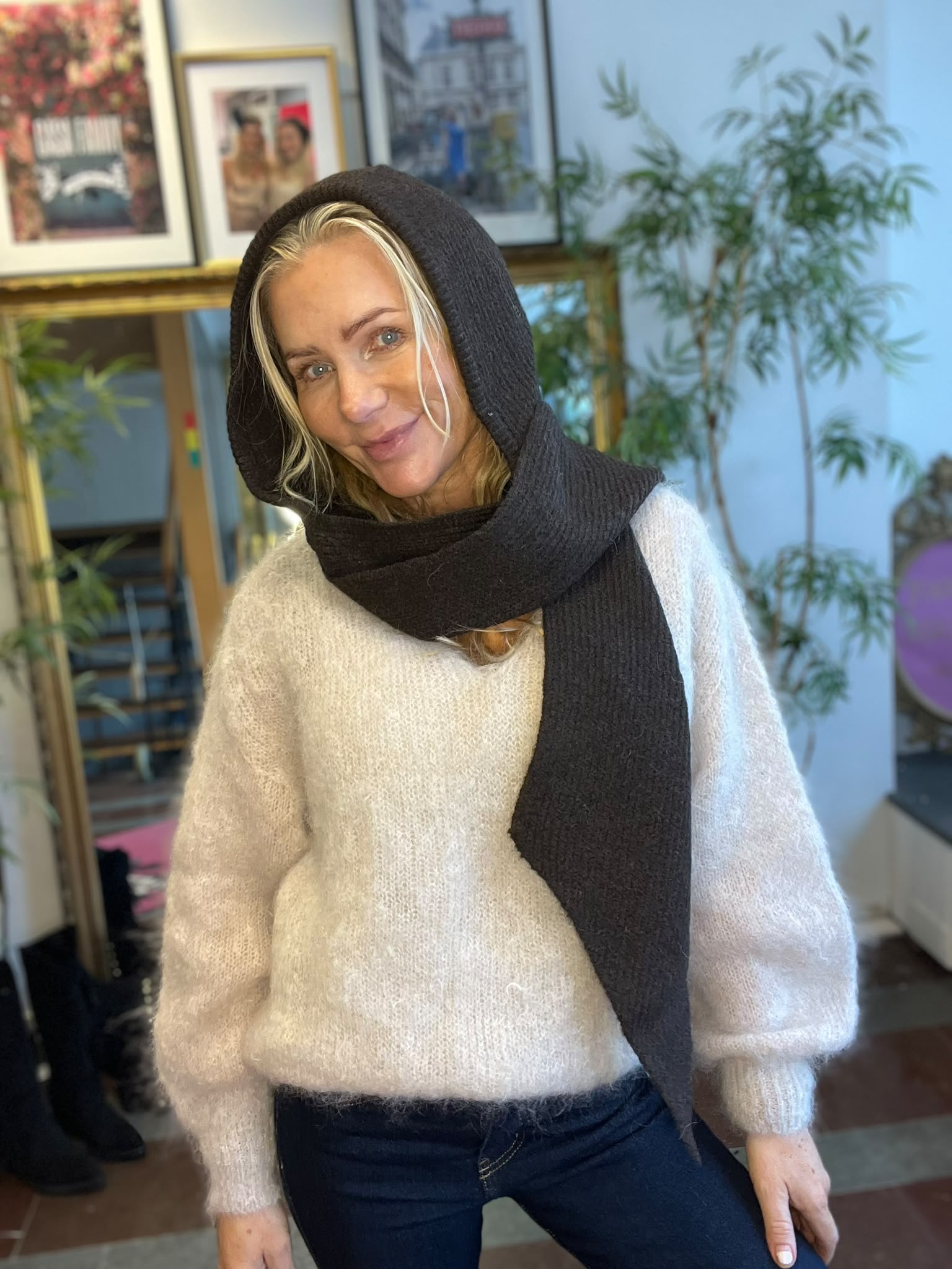 Kvinna med blont hår ler mot kameran och lutar sig lätt framåt, med en mörk stickad sjal som huva och en ljus stickad tröja. Inomhusmiljö med inramade bilder och en grön växt i bakgrunden.